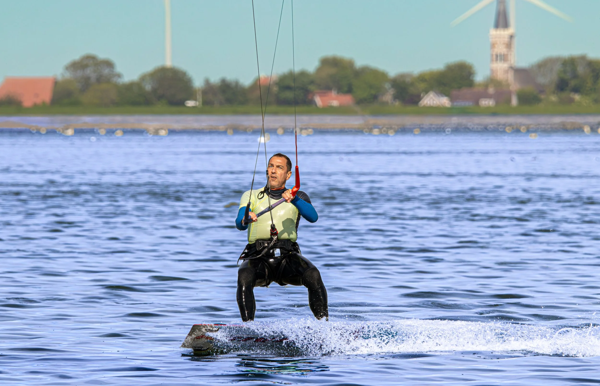 Man die kite surft op het water met een dorp en windmolen op de achtergrond.