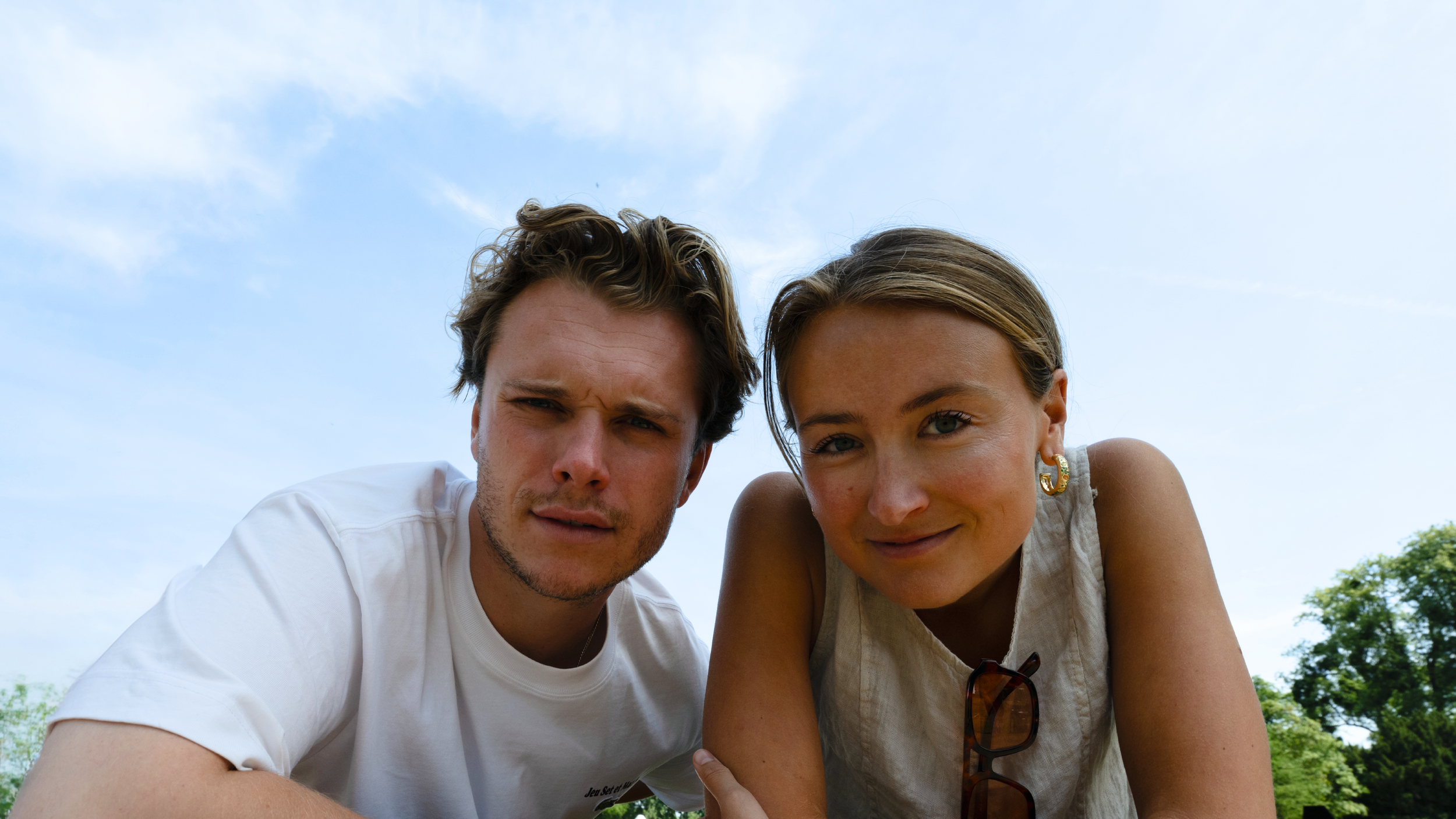 Close-up of a young man with short brown hair and a young woman with light brown hair and sunglasses hanging from her shirt, both smiling and looking at the camera against a blue sky and greenery.