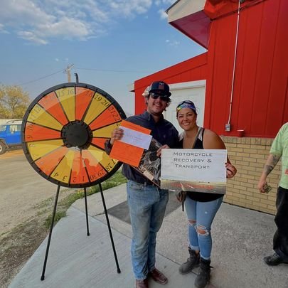 Two people smiling, holding a large check and certificate, standing in front of a prize wheel outside a red building, celebrating motorcycle recovery and transport.