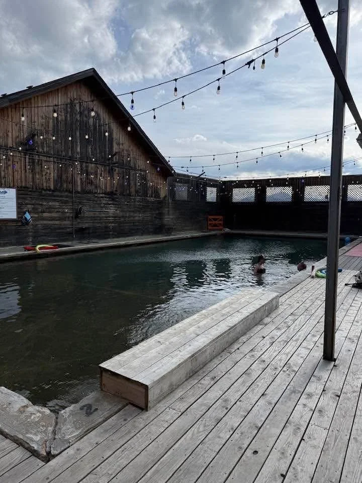 An outdoor swimming pool with a wooden deck and a rustic wooden building in the background, decorated with string lights. Several people are in the pool.