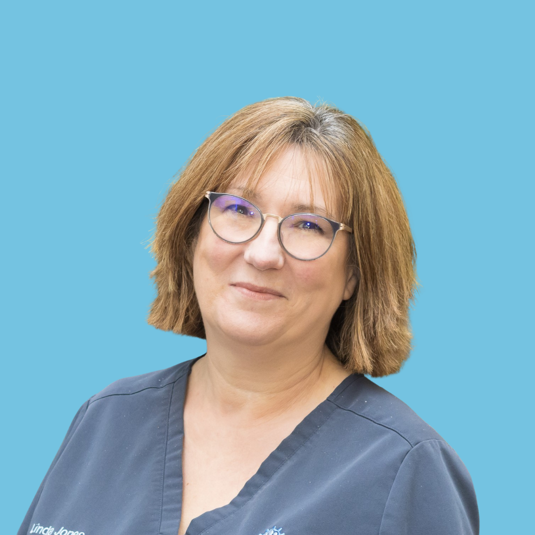 A woman with shoulder-length brown hair, wearing glasses, a black shirt, and a colorful patterned jacket, smiling against a light blue background.