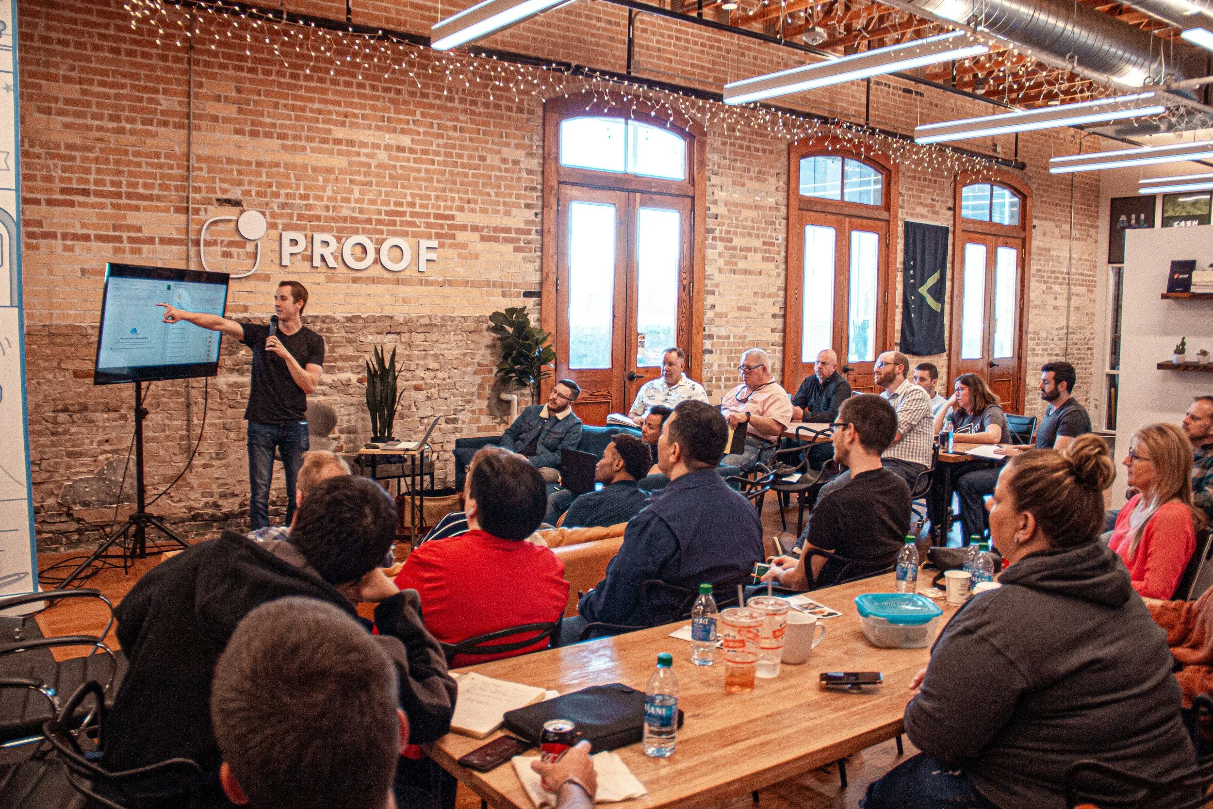 A man in a black shirt giving a presentation to a group of people seated in a loft-style room with exposed brick walls and large windows, with a large monitor displaying slides and string lights overhead.