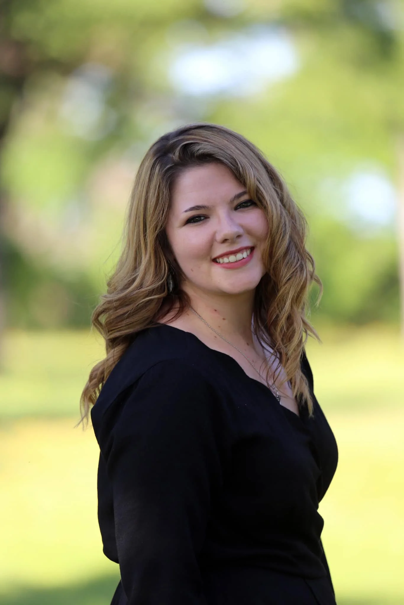 A young realtor with long wavy brown posing in front of nature in Central Texas.