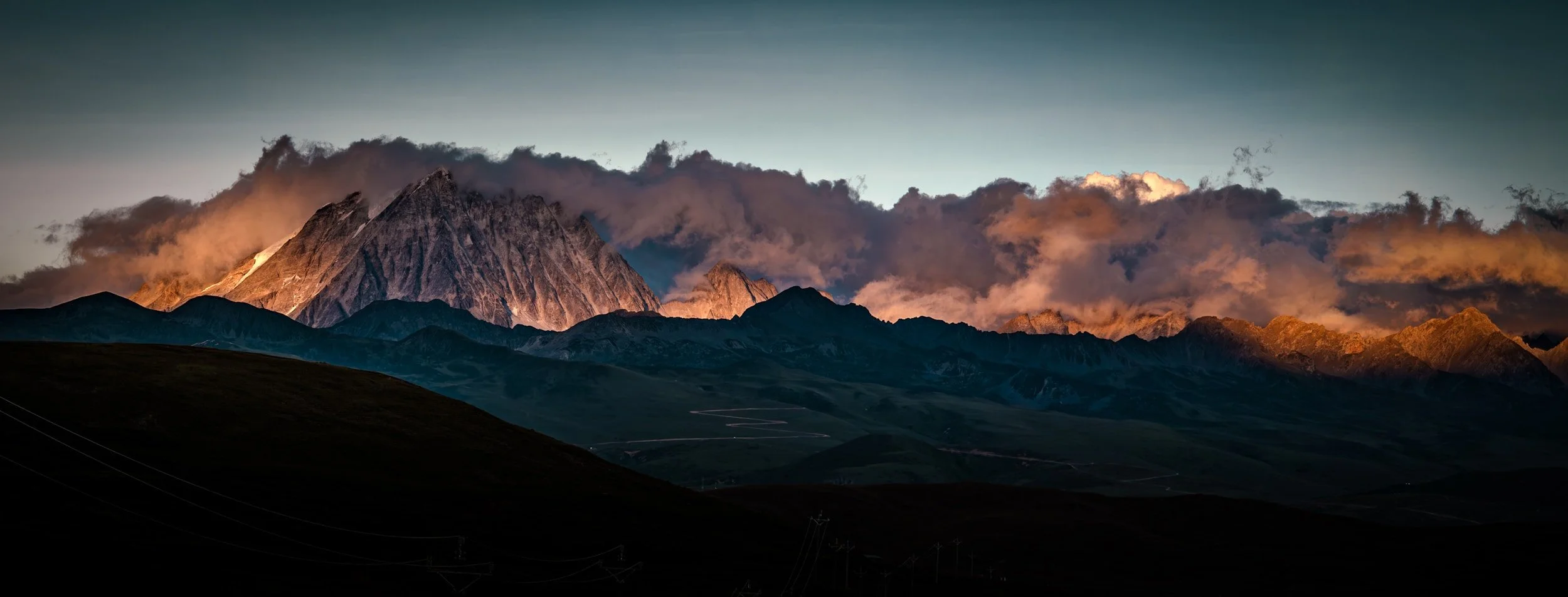 Dramatic winter mountain panorama at sunset with warm golden light on rugged peaks and low clouds, conveying diligence, resilience, emotional strength, and nature-rooted wellbeing.