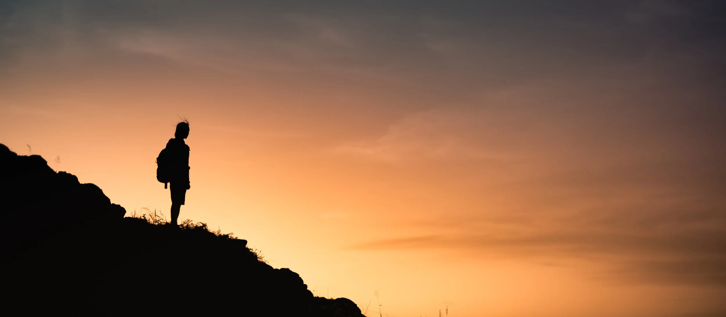 A person standing on a mountain ridge at sunrise, silhouetted against a warm sky, symbolizing reflection, grounding, and reconnecting with oneself.