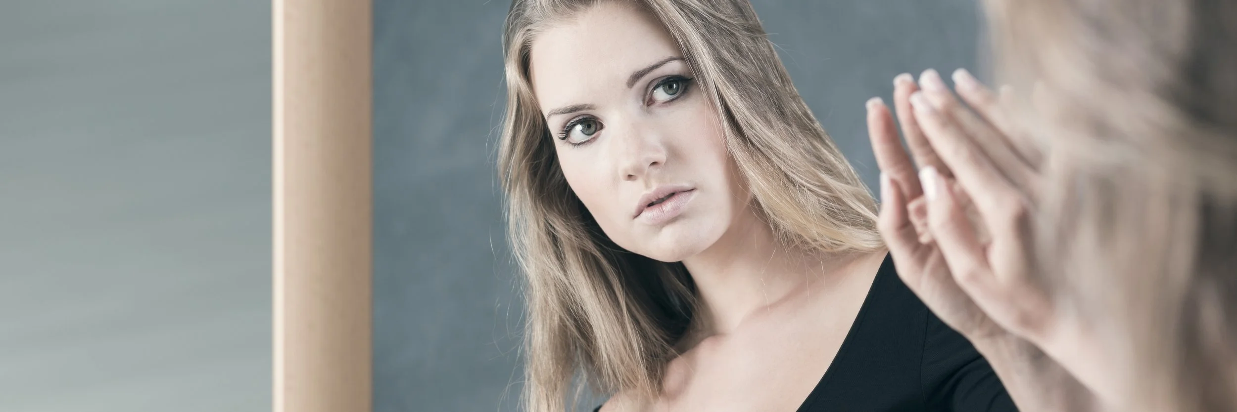 A young woman looking at herself in a mirror with a thoughtful, uncertain expression, symbolizing the emotional complexity of body image.