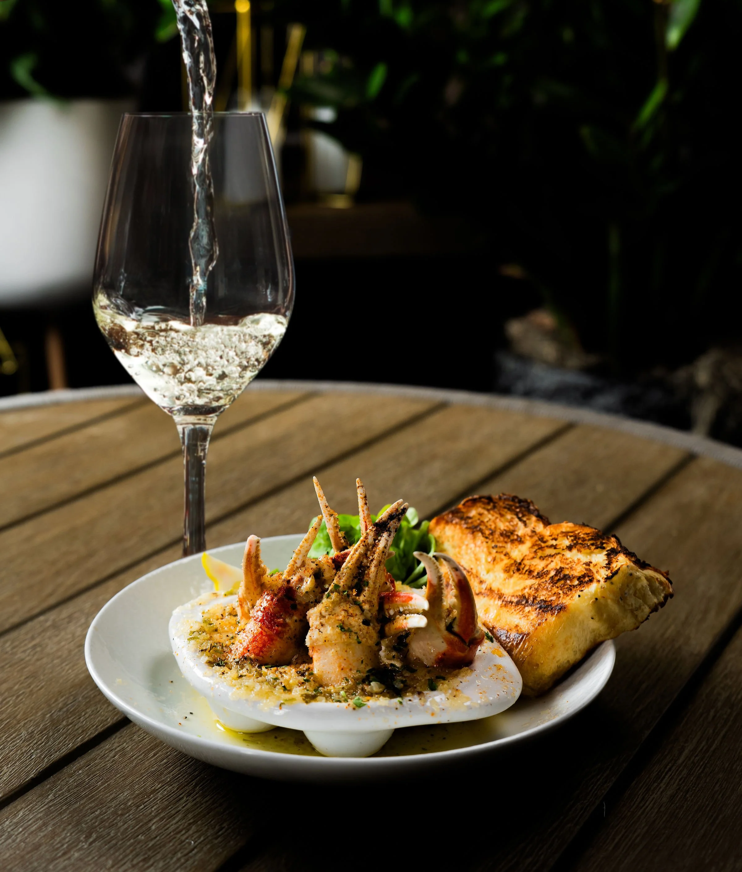 White wine being poured into a glass beside a plate of snow crab escargot and toasted bread, set on an outdoor dining table with greenery in the background.