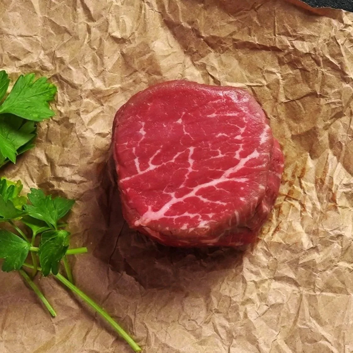 Close-up of a raw filet mignon steak with visible marbling placed on brown butcher paper, garnished with fresh parsley.
