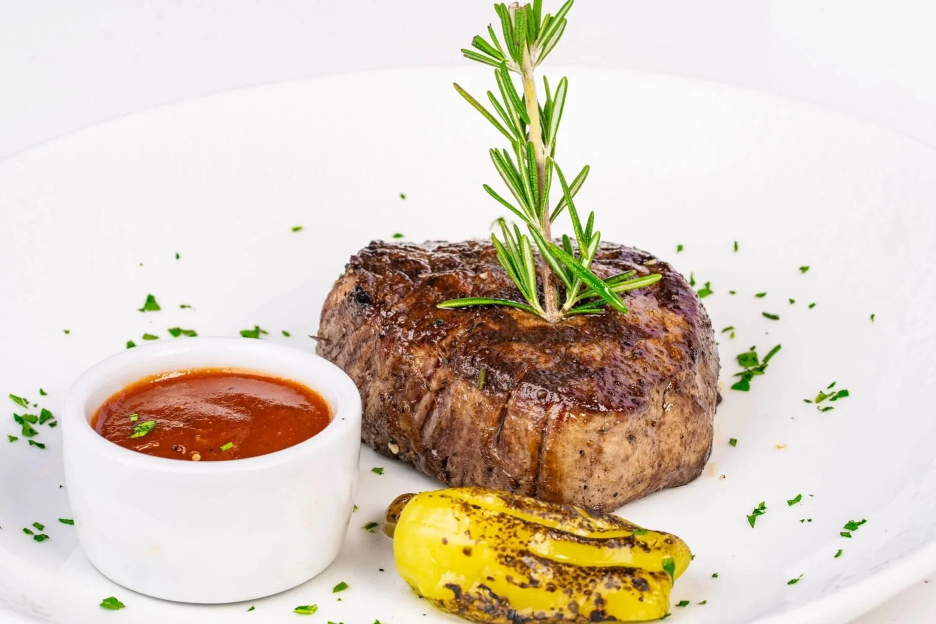 Close-up image of a grilled filet mignon topped with fresh rosemary and served with steak sauce and a charred pepper garnish. The dish is plated on a white plate with fresh herbs, highlighting Catch & Cut’s premium steak offerings.