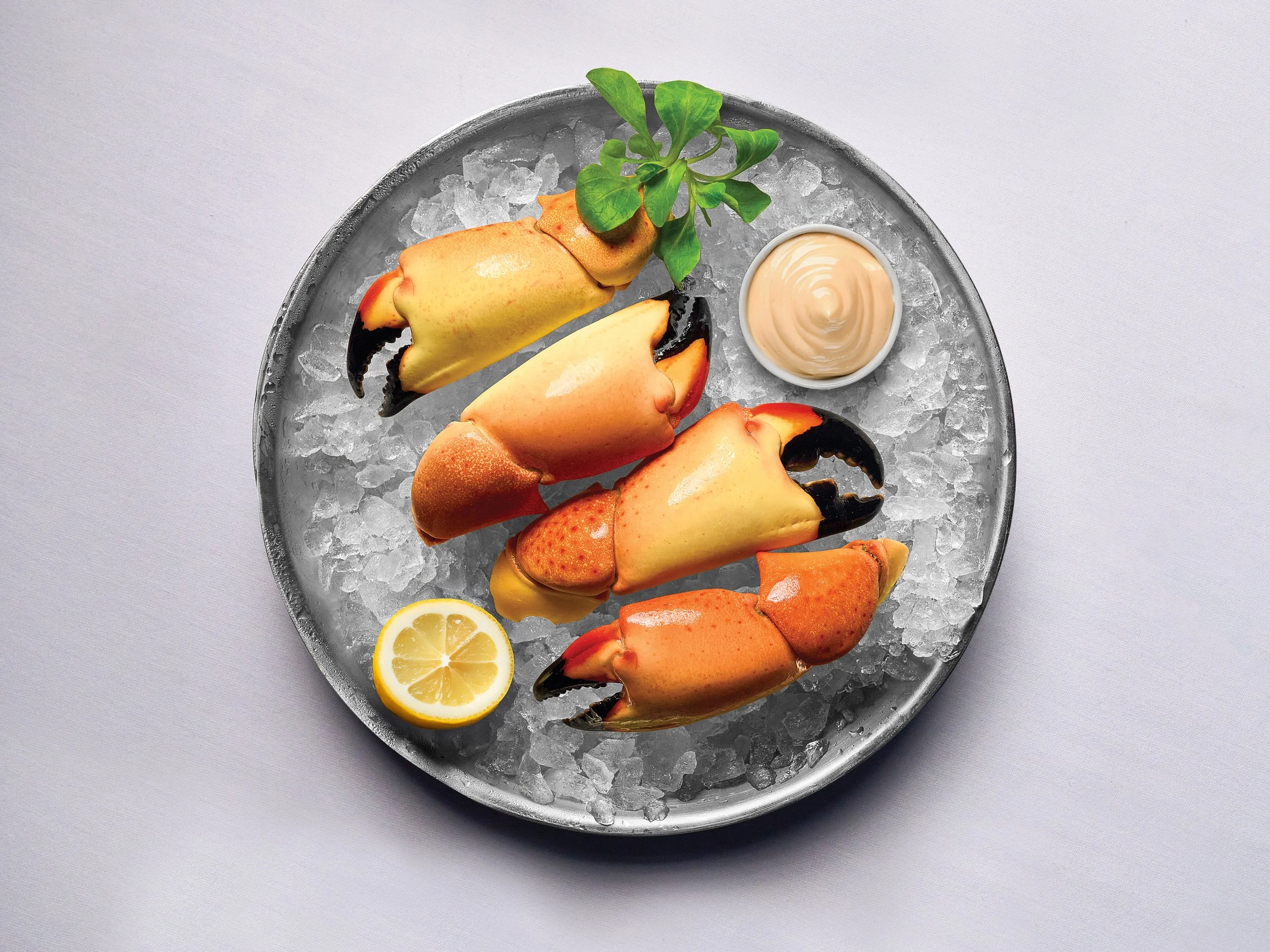 Four Stone Crab Claws on a bed of ice, garnished with lemon and a small bowl of Catch & Cut Signature Mustard sauce, on a round silver tray.