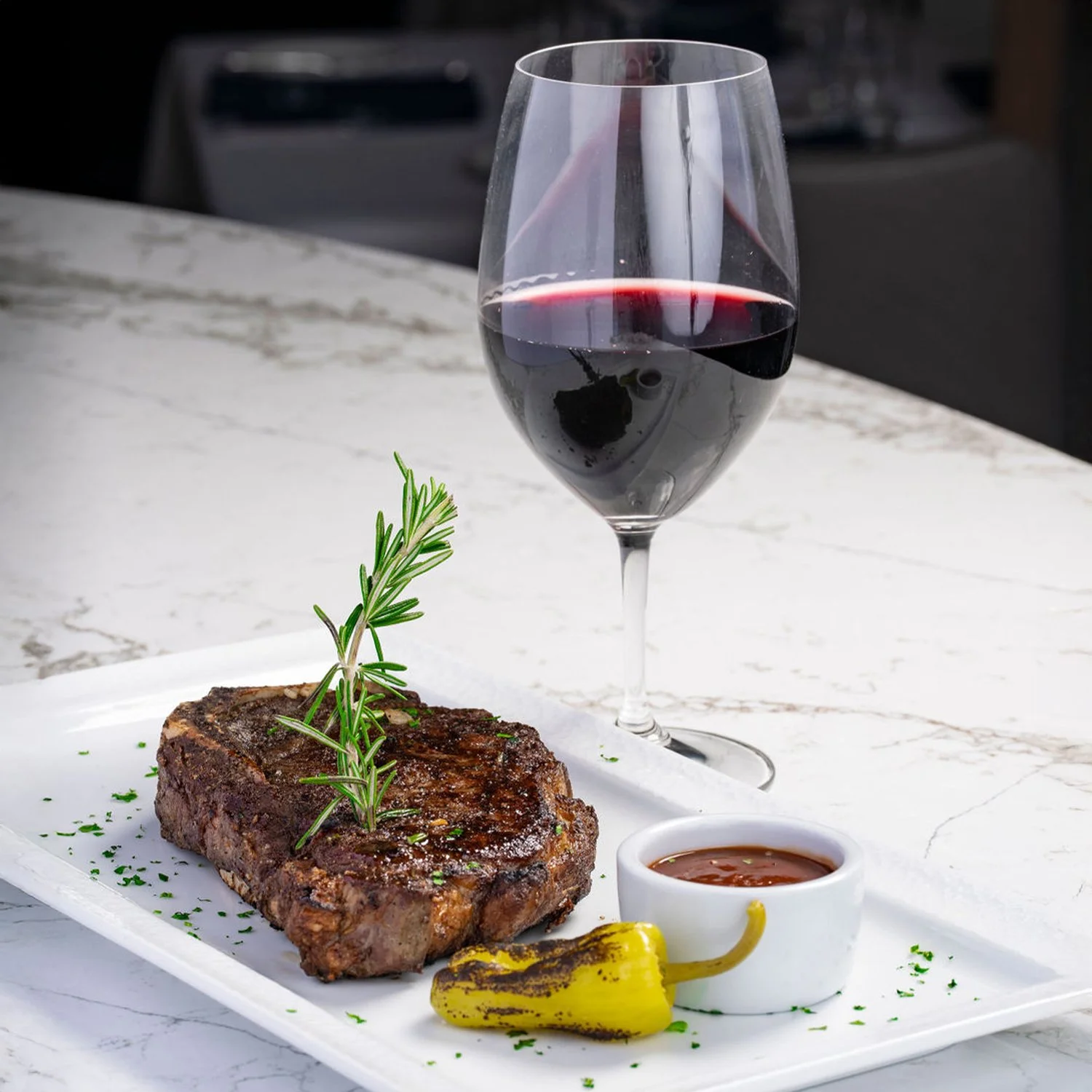 A glass of red wine, a USDA Prime Rib Eye garnished with rosemary, a roasted yellow pepper, and a small cup of sauce on a white rectangular plate, set on a marble table.