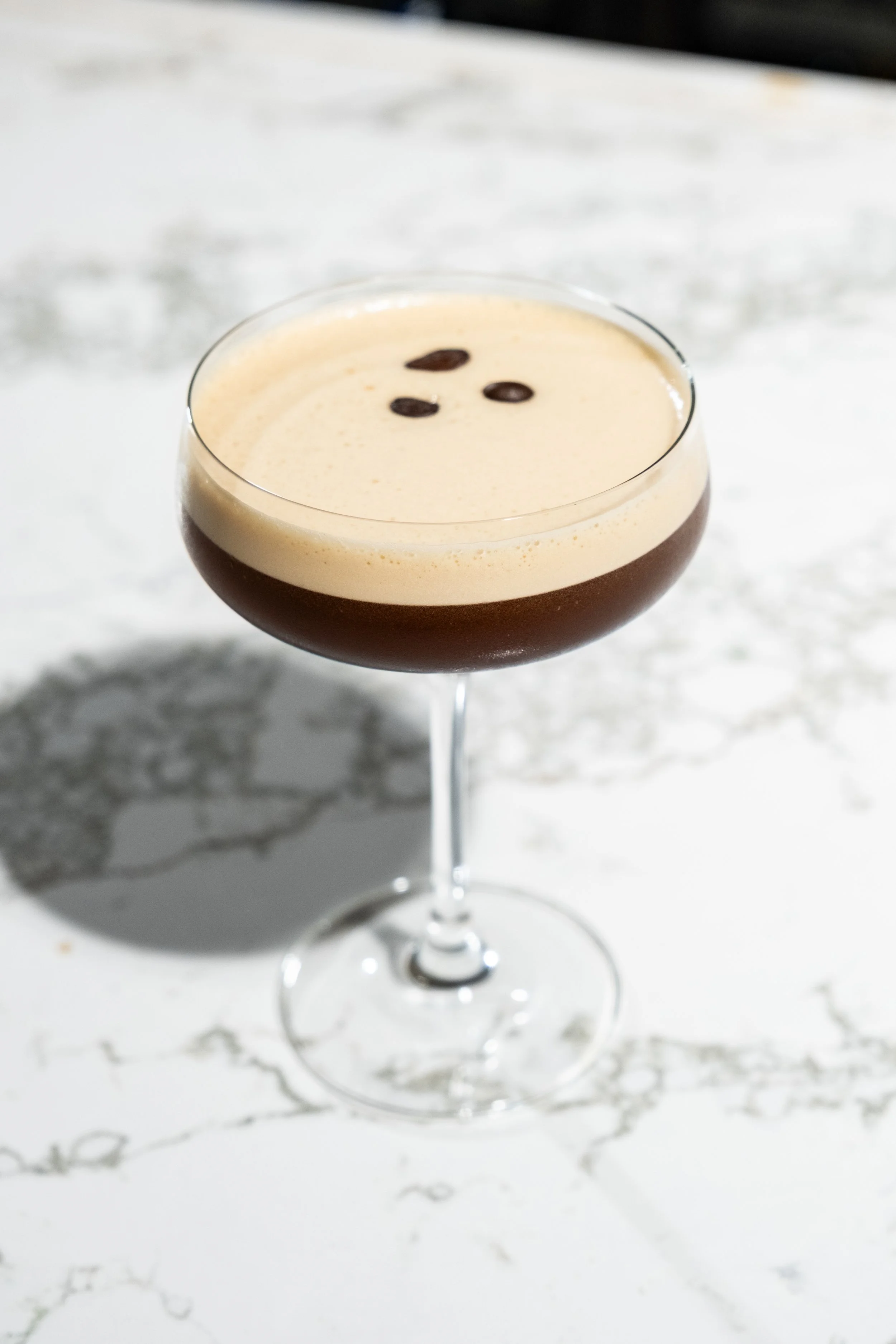Catch & Cut's signature espresso martini cocktail in a coupe glass with a frothy top and three coffee beans garnishing it, placed on a white marble surface.