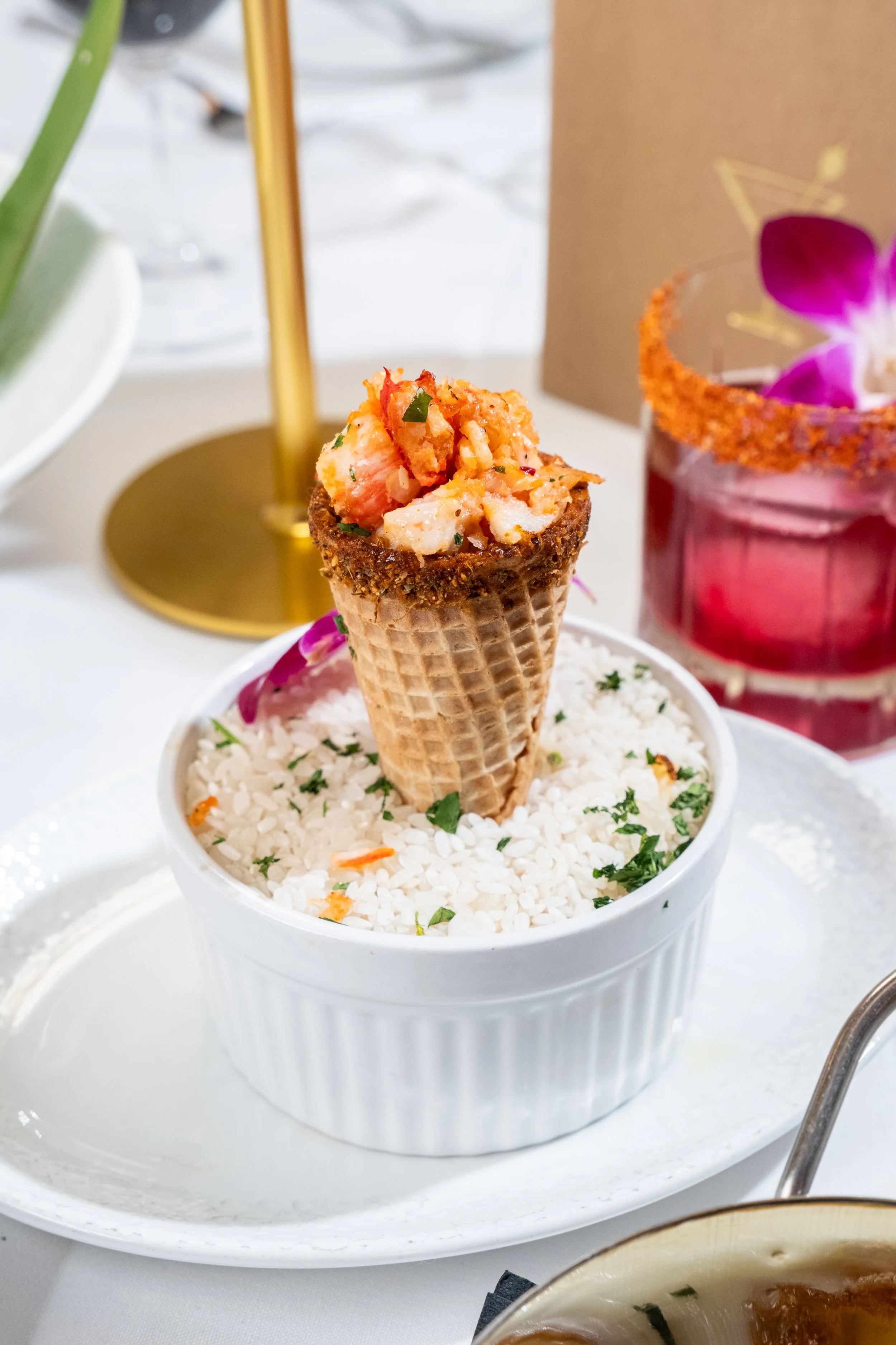 Lobster cone filled with lobster meat, served upright in a crisp waffle cone over seasoned rice, garnished with fresh herbs and edible flowers on a white plate.