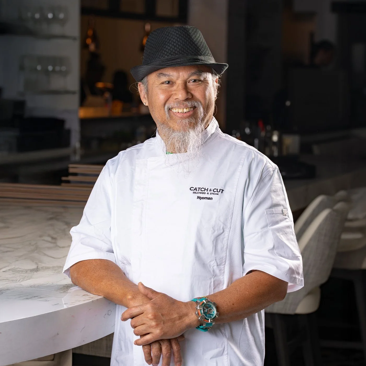 Catch & Cut Head Sushi Chef, Inyoman Atmaja, smiling with a beard and gray hair wearing a white chef's coat and a black fedora hat, standing in the main dining room at Catch & Cut restaurant