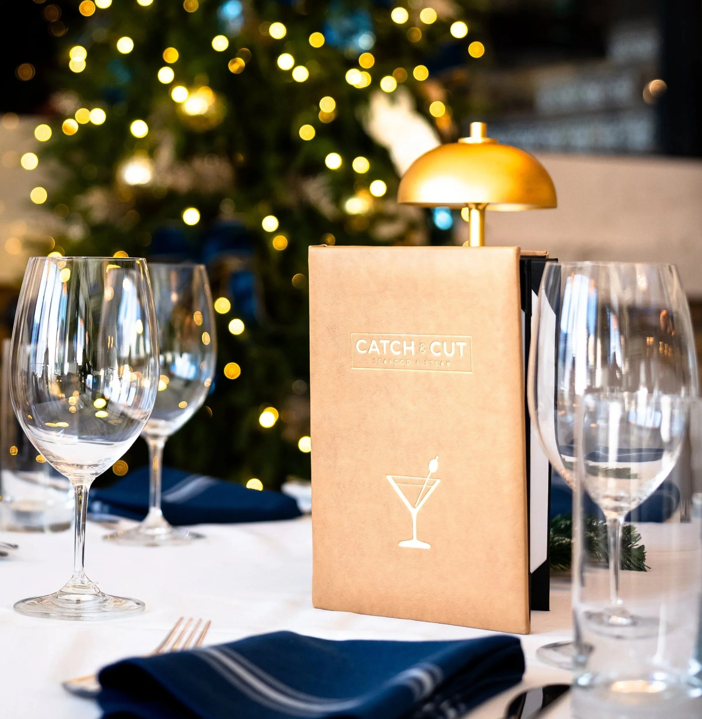 Catch & Cut table set for dinner with four wine glasses, a menu from Catch & Cut, and a gold lamp, with Christmas lights and a decorated Christmas tree in the background.