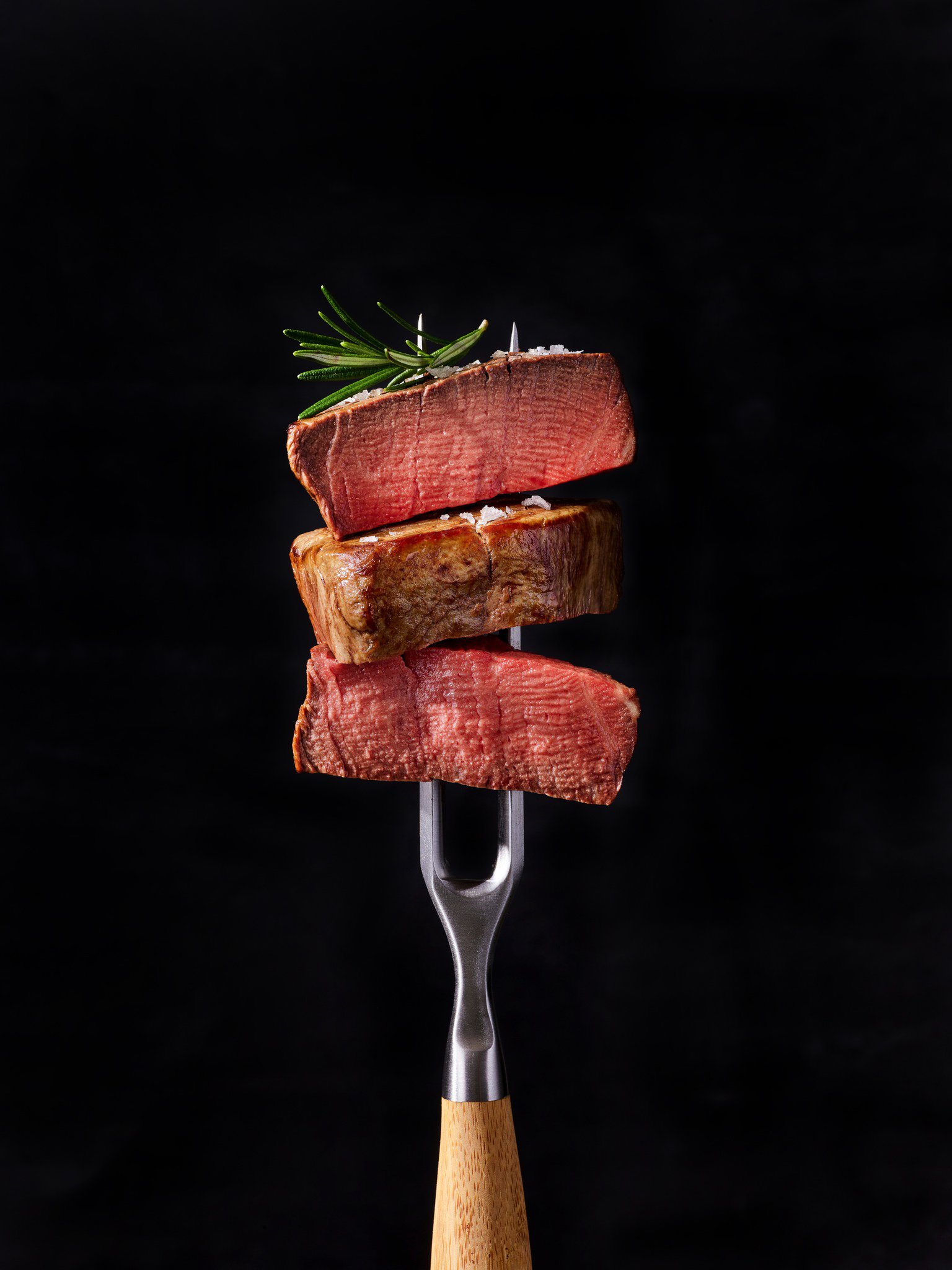 Three slices of medium-rare steak stacked on a fork with a sprig of rosemary on top against a black background.