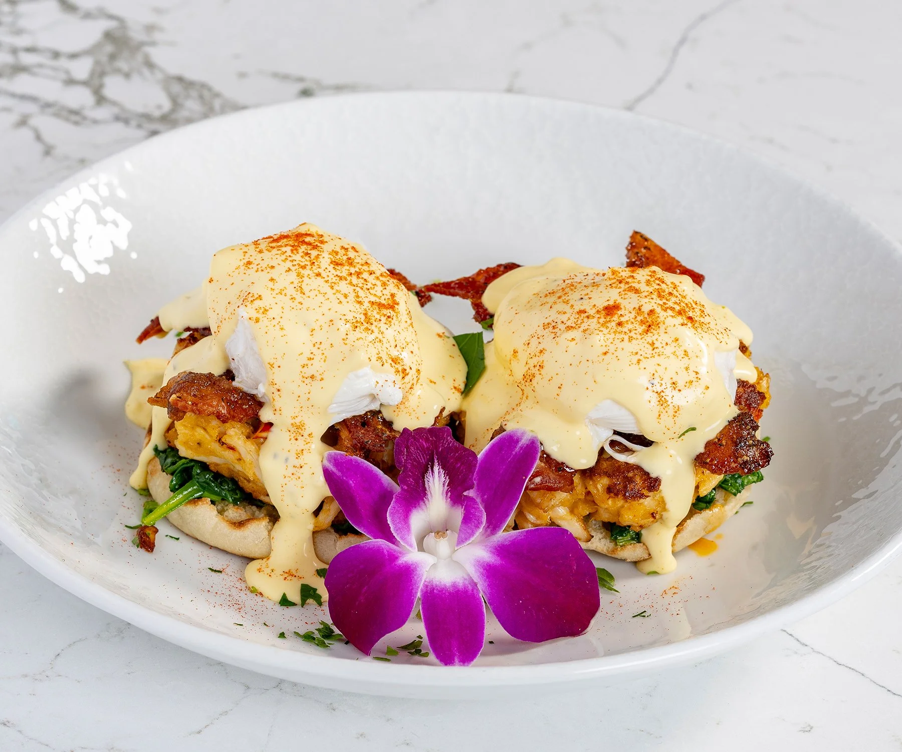 Two eggs Benedict with bacon on toasted English muffins and a purple orchid flower on a white plate.
