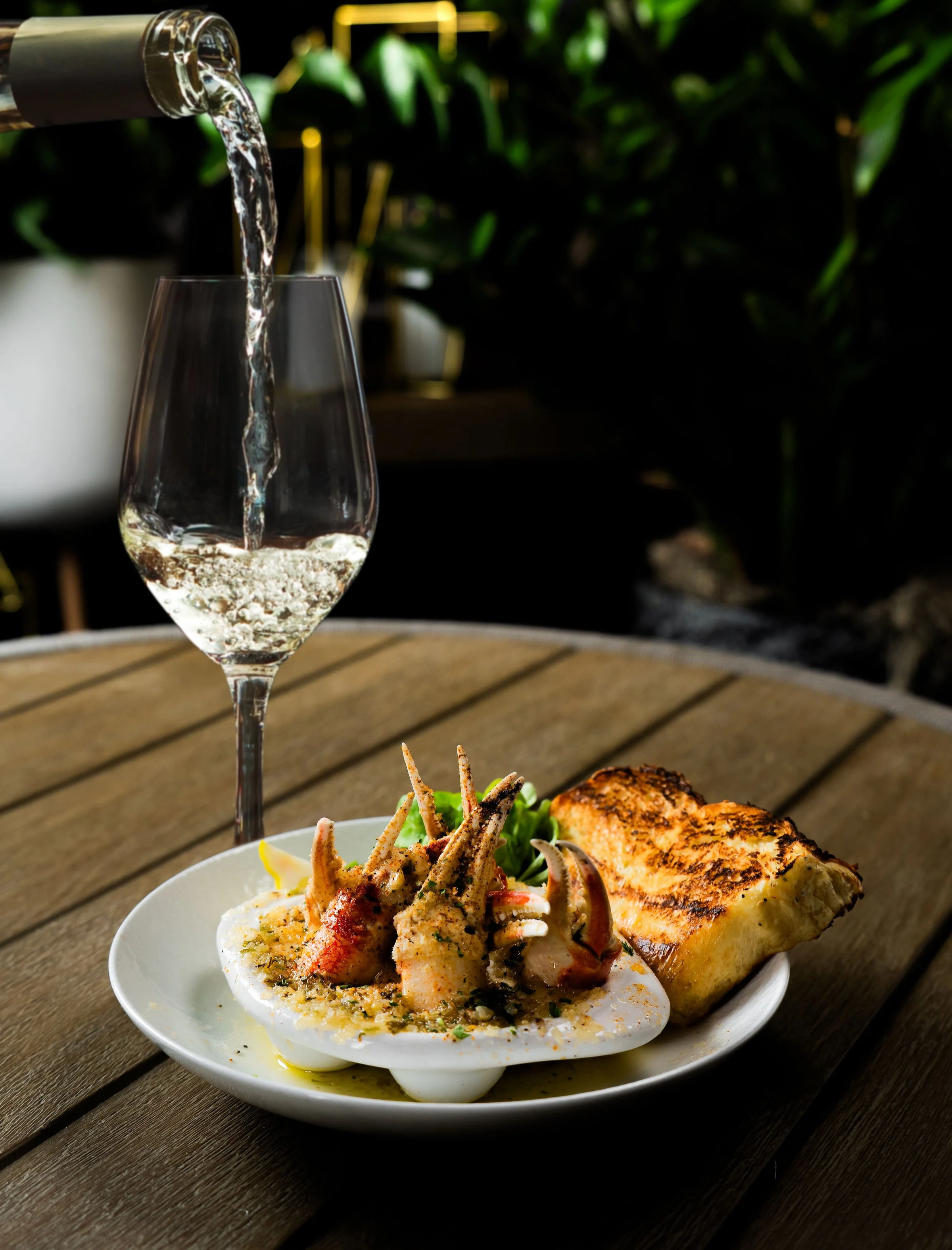 White wine being poured into a glass beside a plate of snow crab escargot and toasted bread, set on an outdoor dining table with greenery in the background.