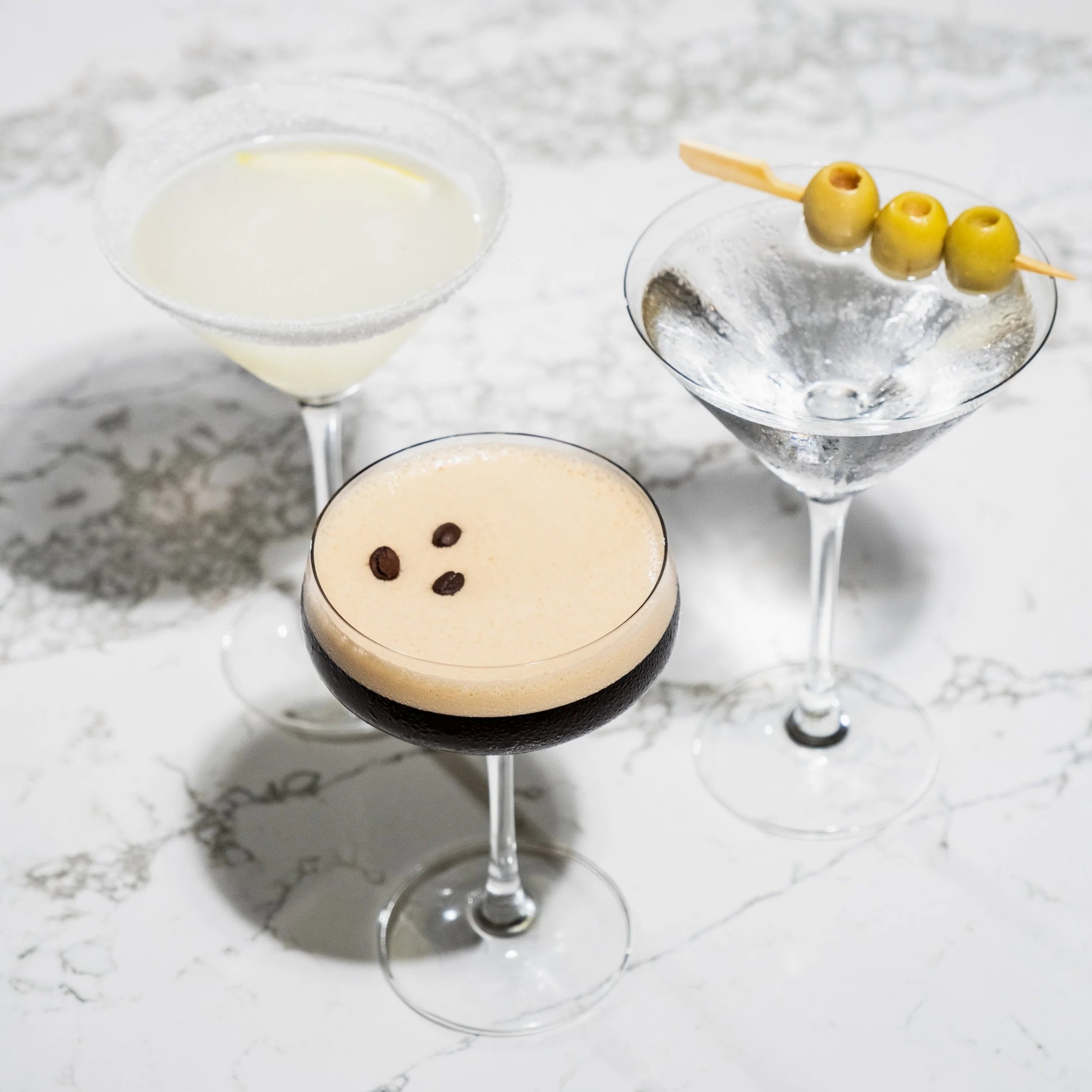 Three Tito’s martinis displayed on a marble table, including an Espresso Martini topped with coffee beans, a Lemon Drop Martini with a sugared rim, and a Classic Vodka Martini garnished with green olives.
