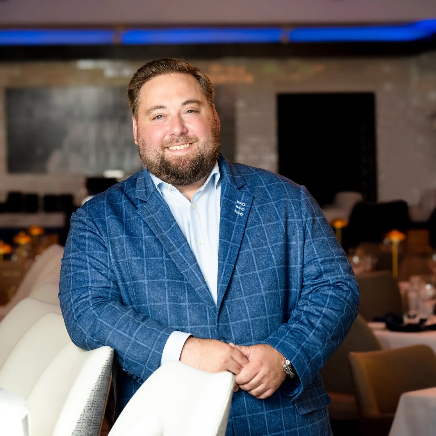 Catch & Cut General Manager, Sherwin Levitis, a man with a beard and brown hair, smiling in a blue checkered suit jacket and white shirt, standing in the main dining room at Catch & Cut restaurant