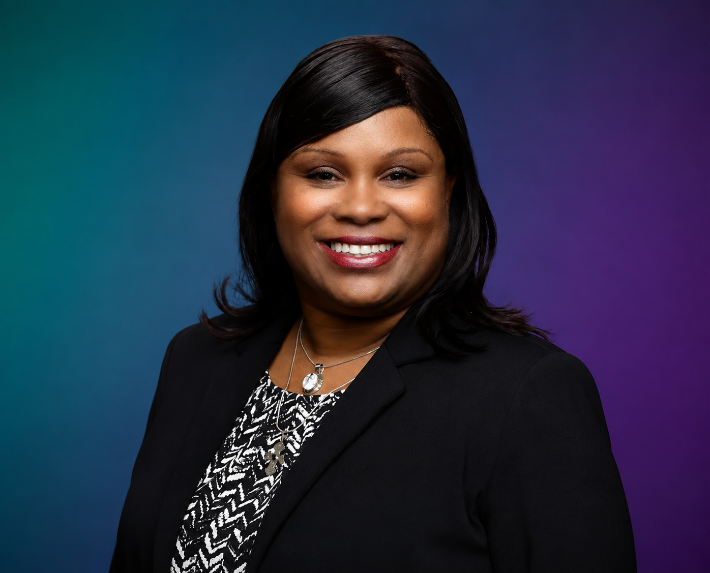 Professional woman with black hair smiling, wearing a black blazer and patterned blouse, against a multicolored gradient background.