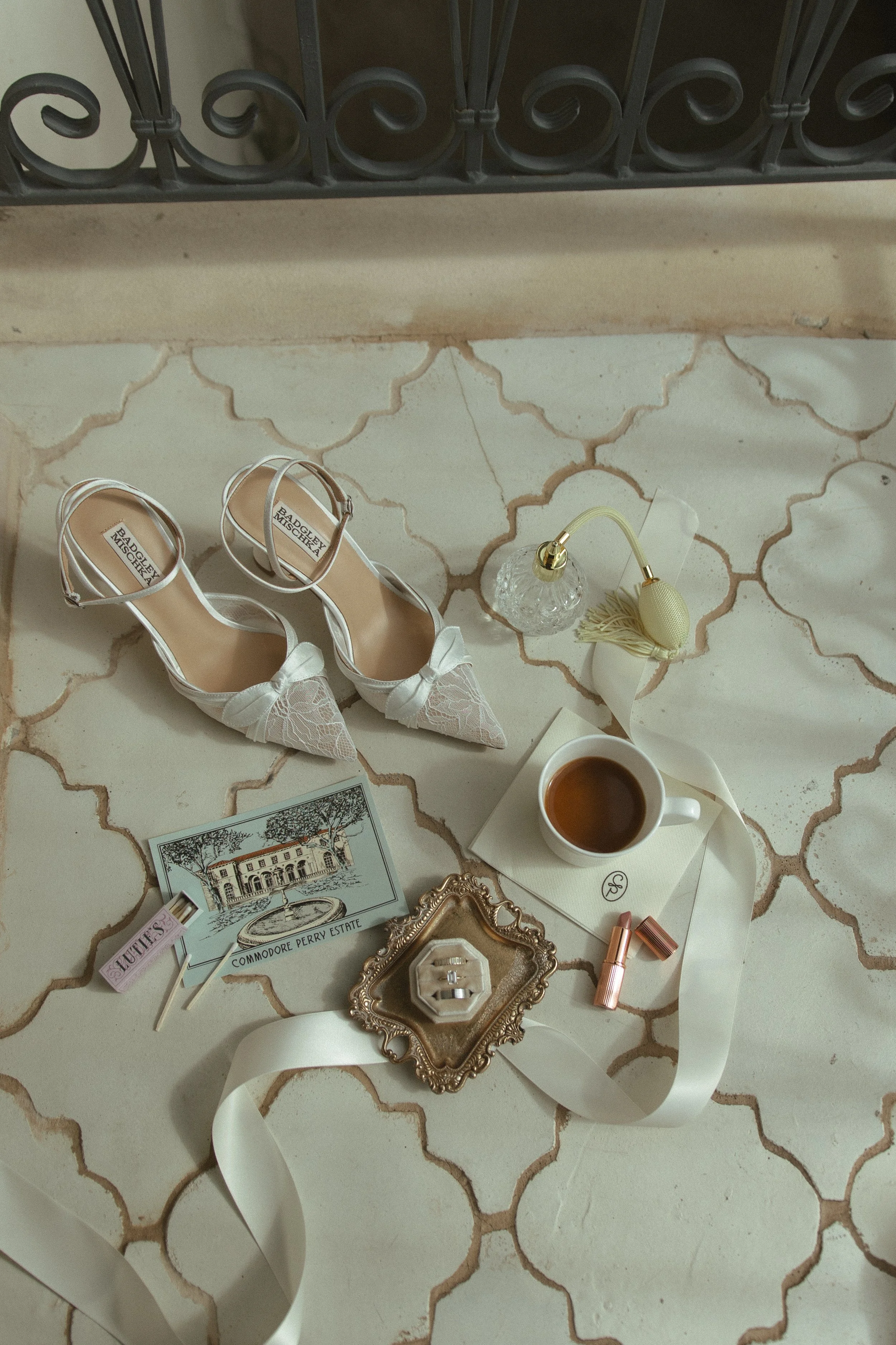 A collection of wedding accessories on a decorative tile floor, including white bridal shoes with bows, a bottle of perfume with a spray nozzle, a cup of coffee on a napkin, a small ornate picture frame, a gold lipstick, a postcard of a historic estate, and a small jewelry box.
