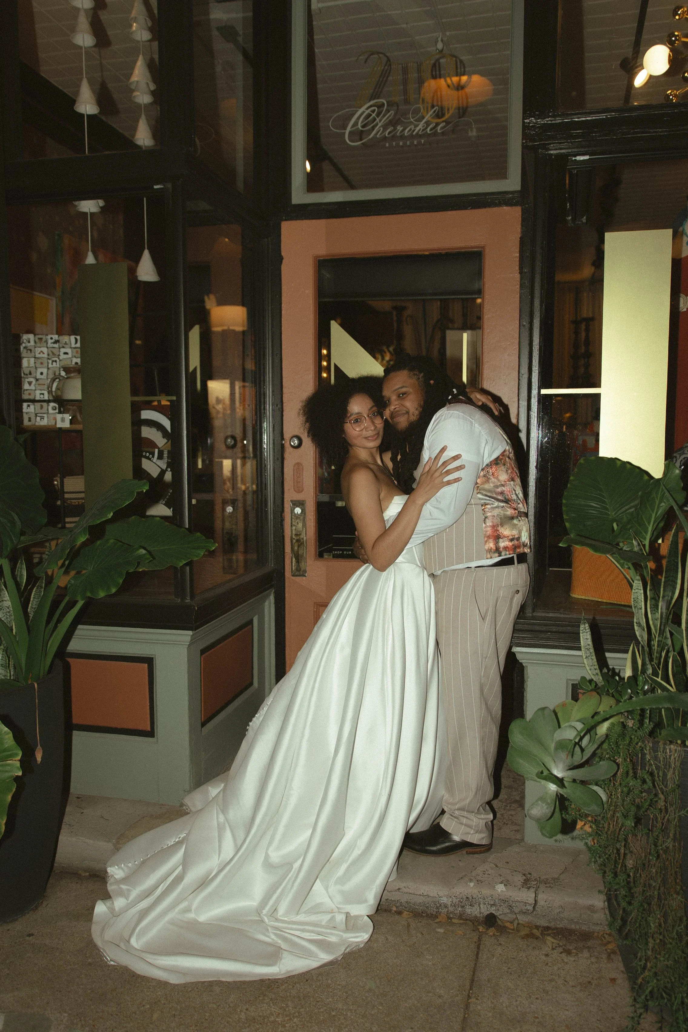 A couple dressed in wedding attire embraces and poses outside a building at night.