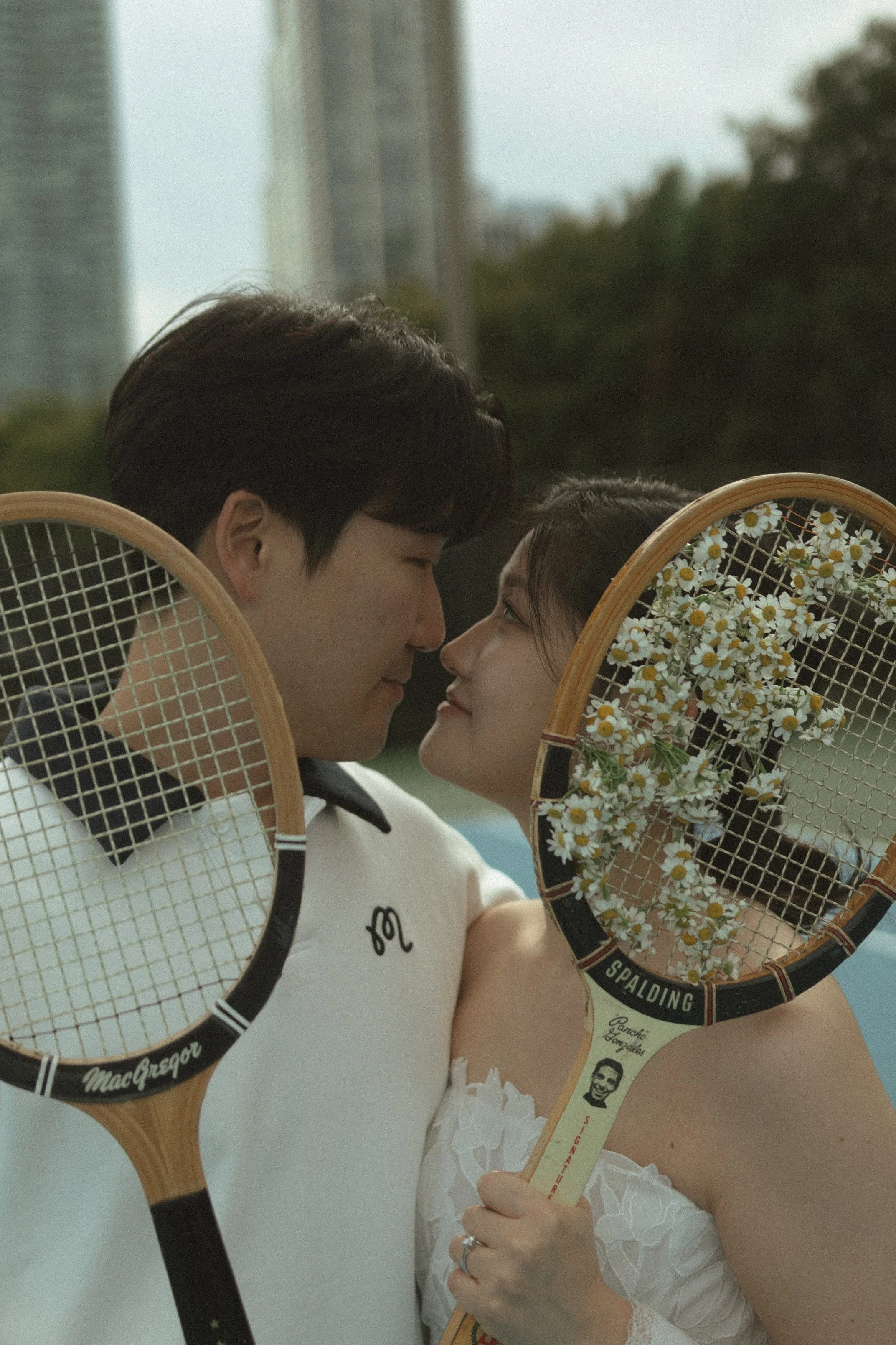 A couple holding tennis rackets close to each other, indicating affection, outside in an urban park with tall buildings in the background.