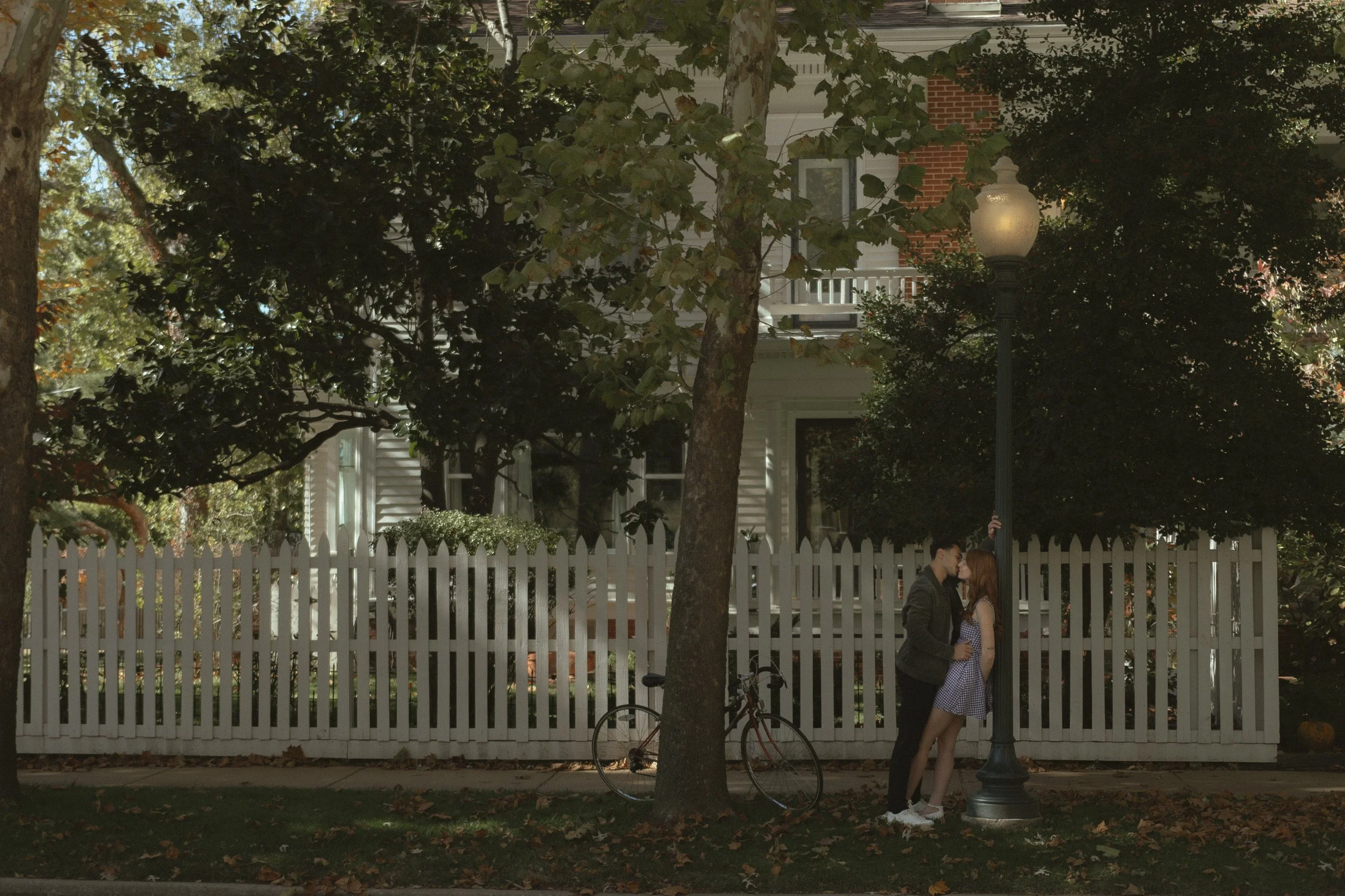 A young couple stands close together, kissing behind a lamppost on a quiet street with a white picket fence and a house in the background. A bicycle leans against a tree nearby, and the ground is covered with fallen leaves, suggesting an autumn setting.