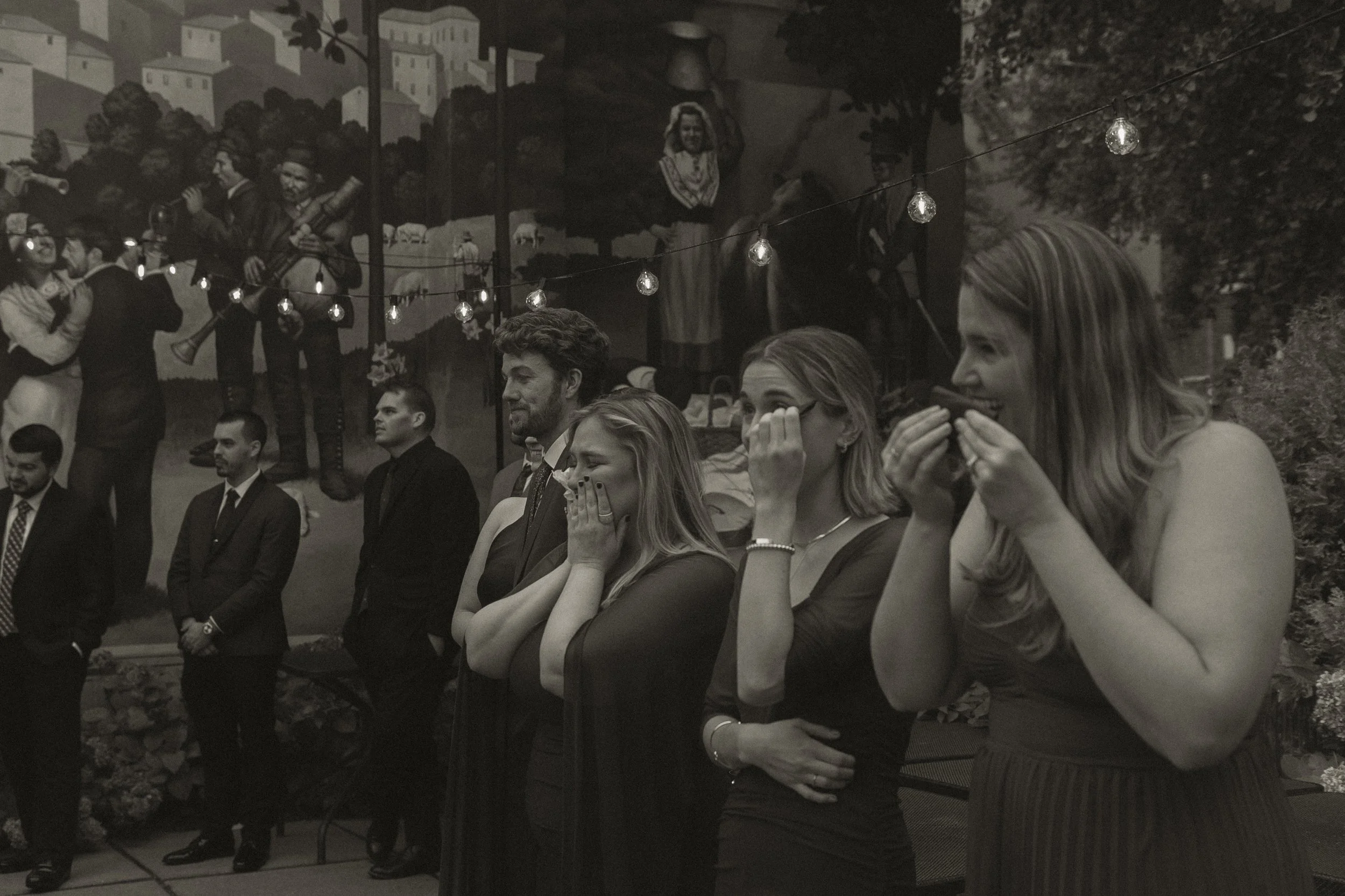 Group of five women standing indoors, reacting with amusement and laughter, with some covering their mouths or covering their eyes.