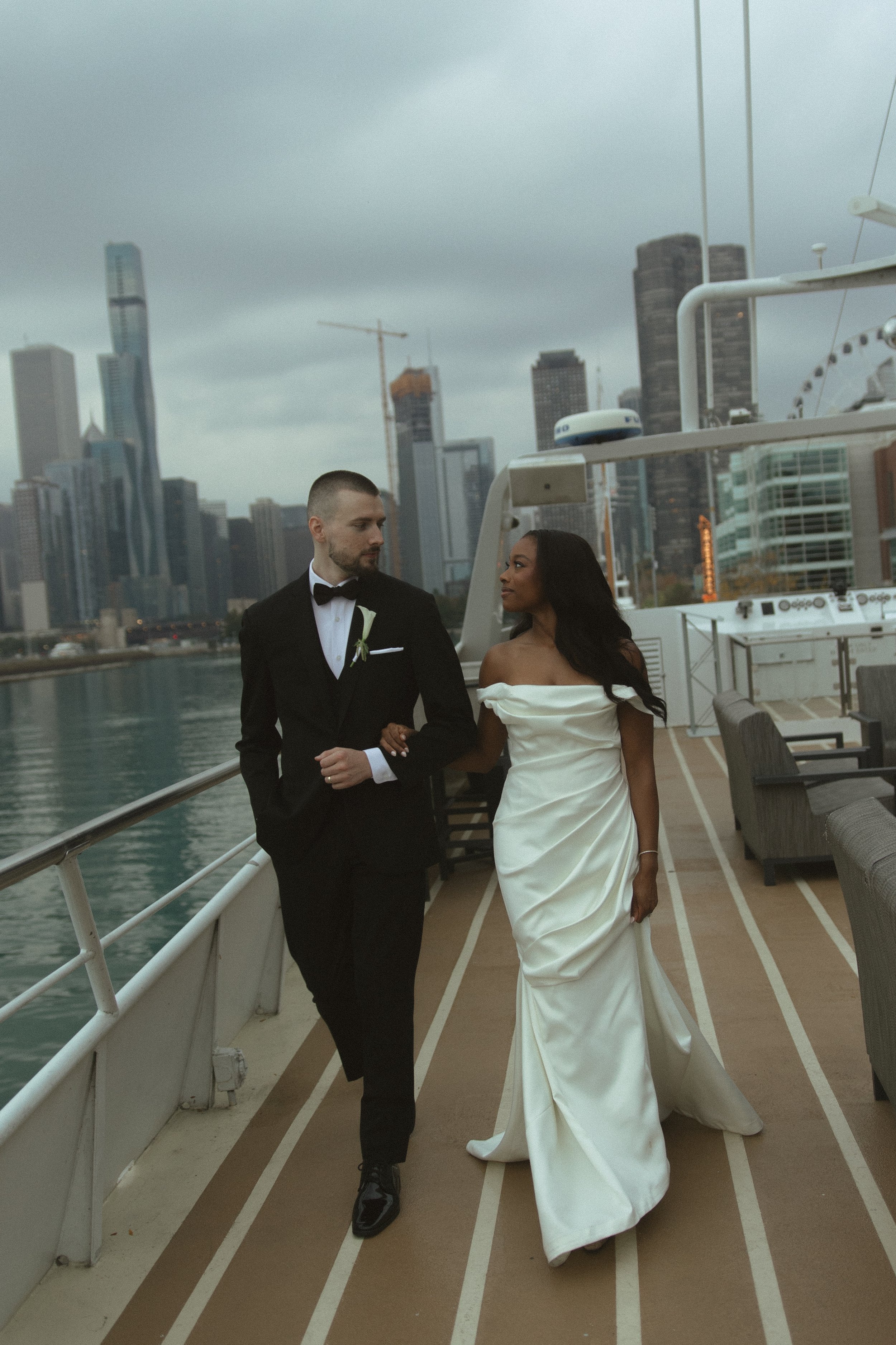 A couple dressed in wedding attire walking on a boat with a city skyline in the background.