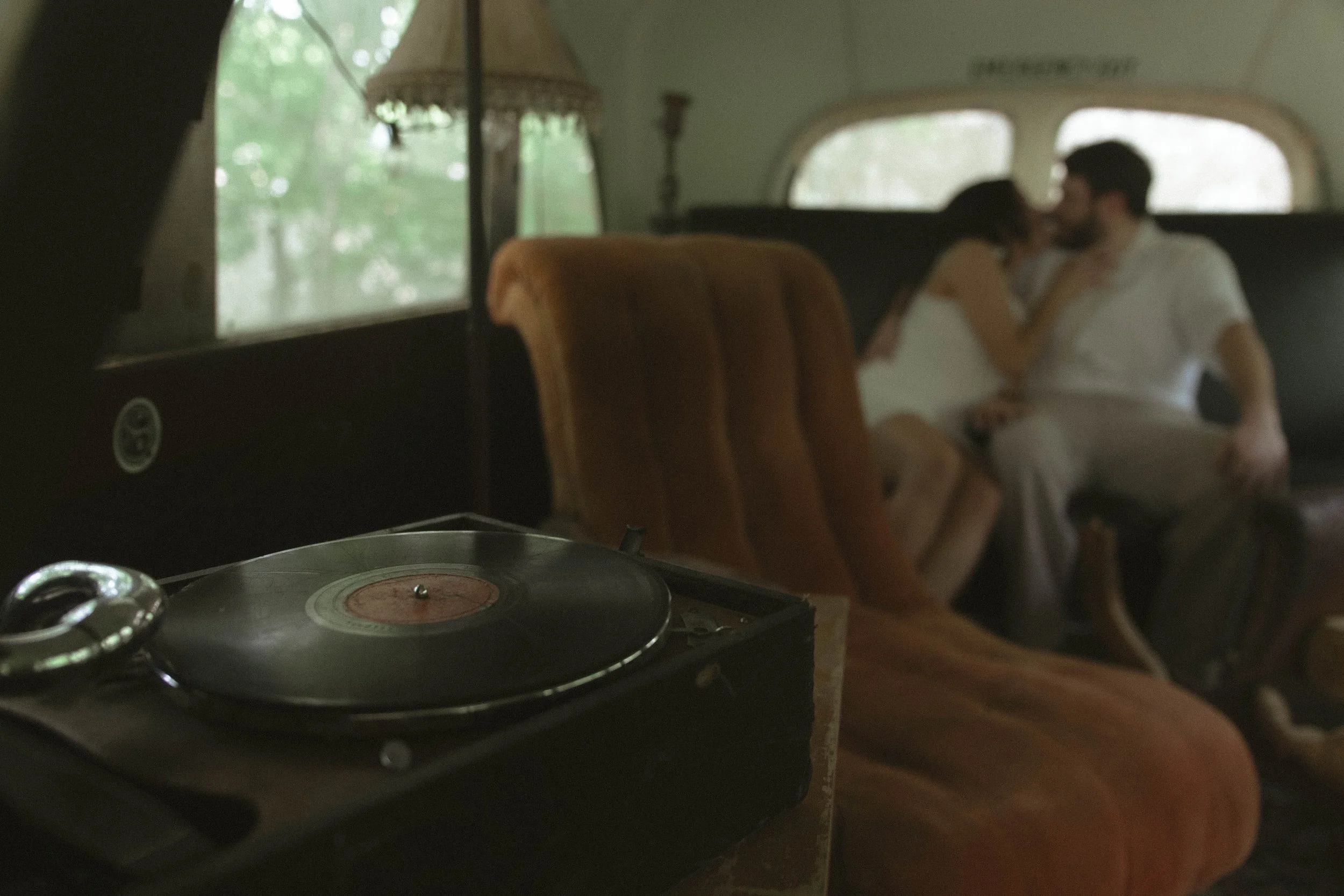 Vintage turntable in the foreground with a blurred couple sitting closely on a couch in the background inside a retro camper or bus, with trees visible outside the window.