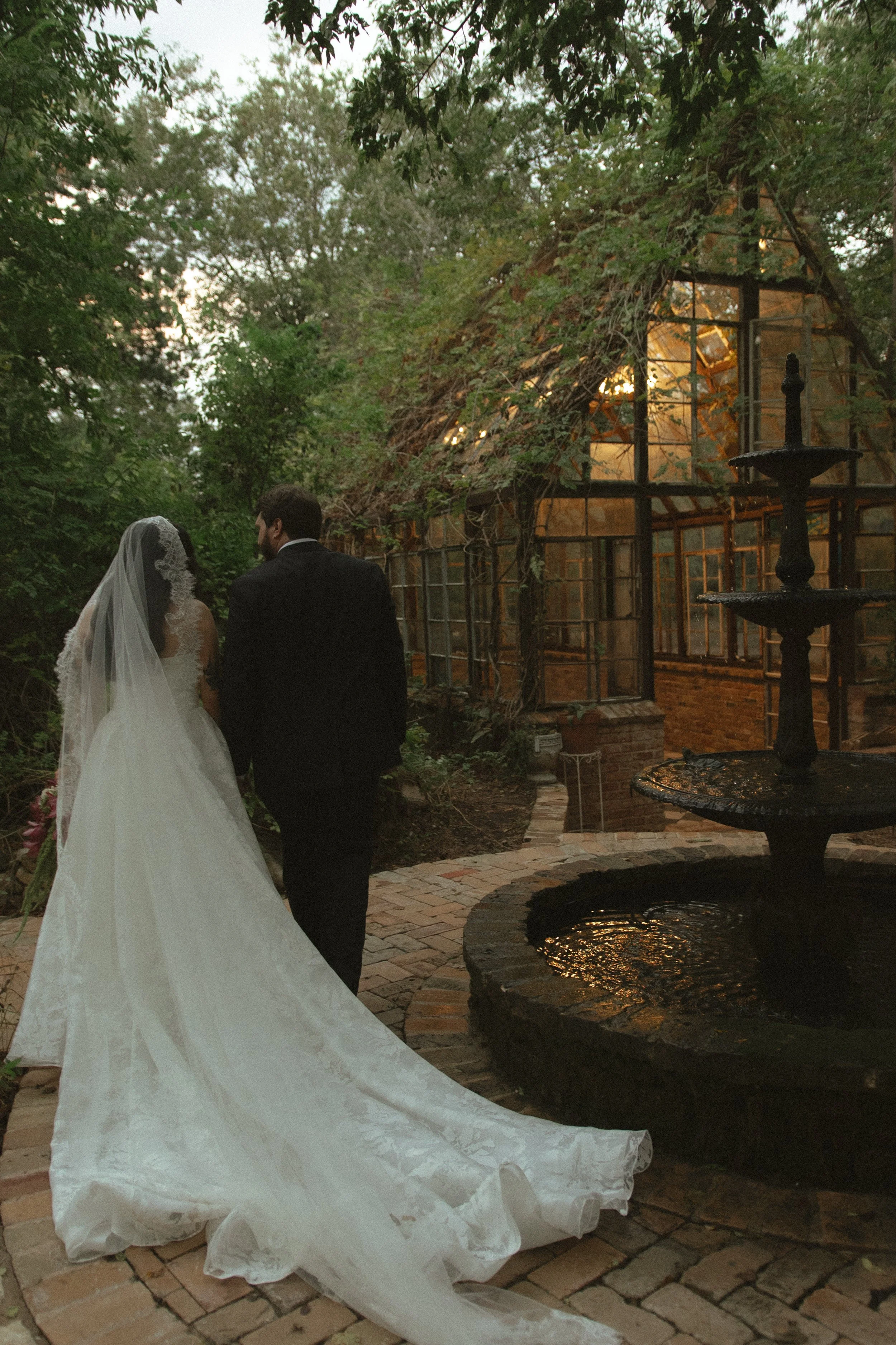 A bride and groom walk together through a garden pathway near an illuminated greenhouse, with a tiered fountain in the foreground and lush trees surrounding them.