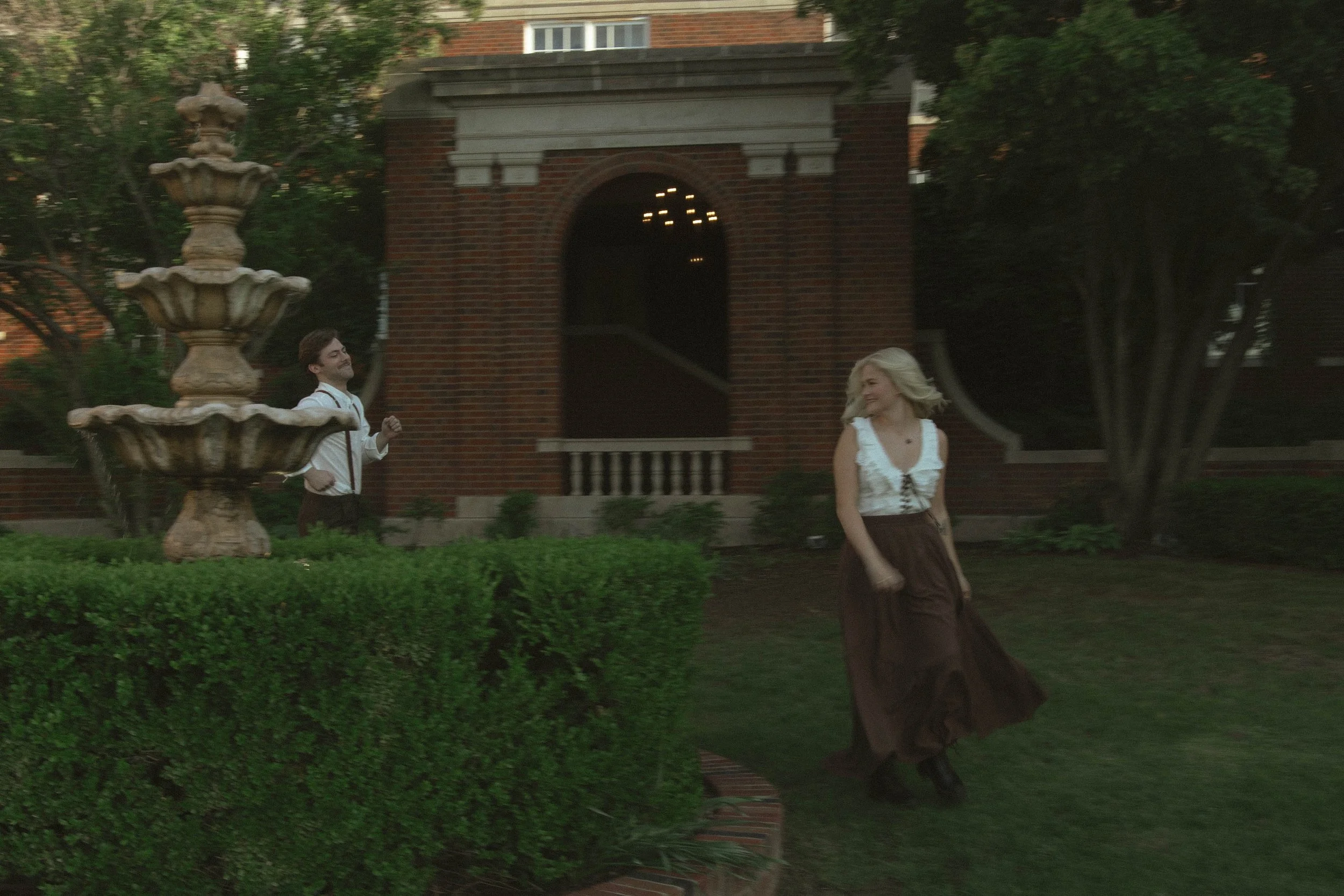 A man and woman are in a garden near a fountain and a brick building, with the woman running or dancing while the man smiles and looks at her.