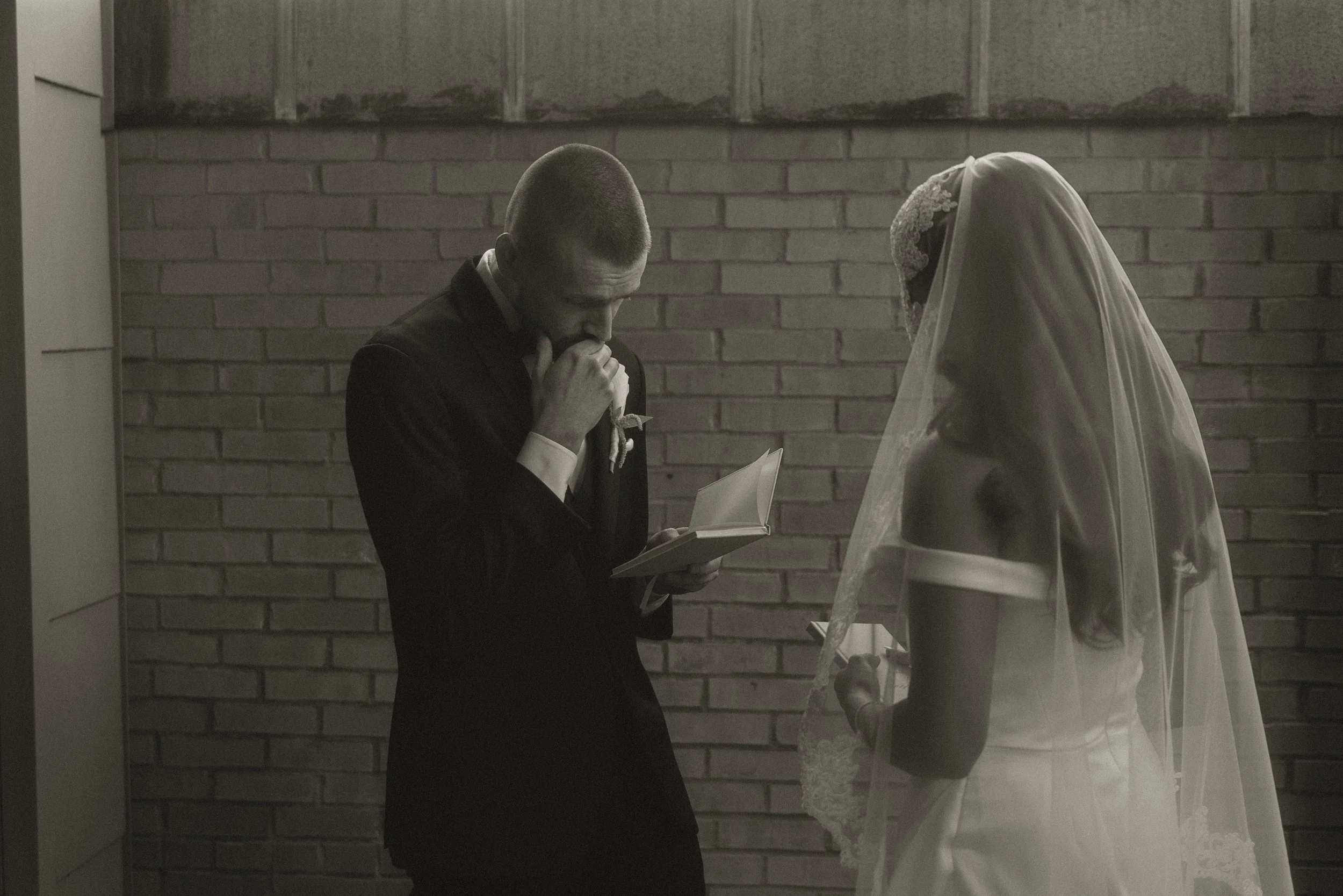 A groom praying with clasped hands while reading vows from a booklet, and a bride in a wedding dress with a veil standing nearby, against a brick wall.