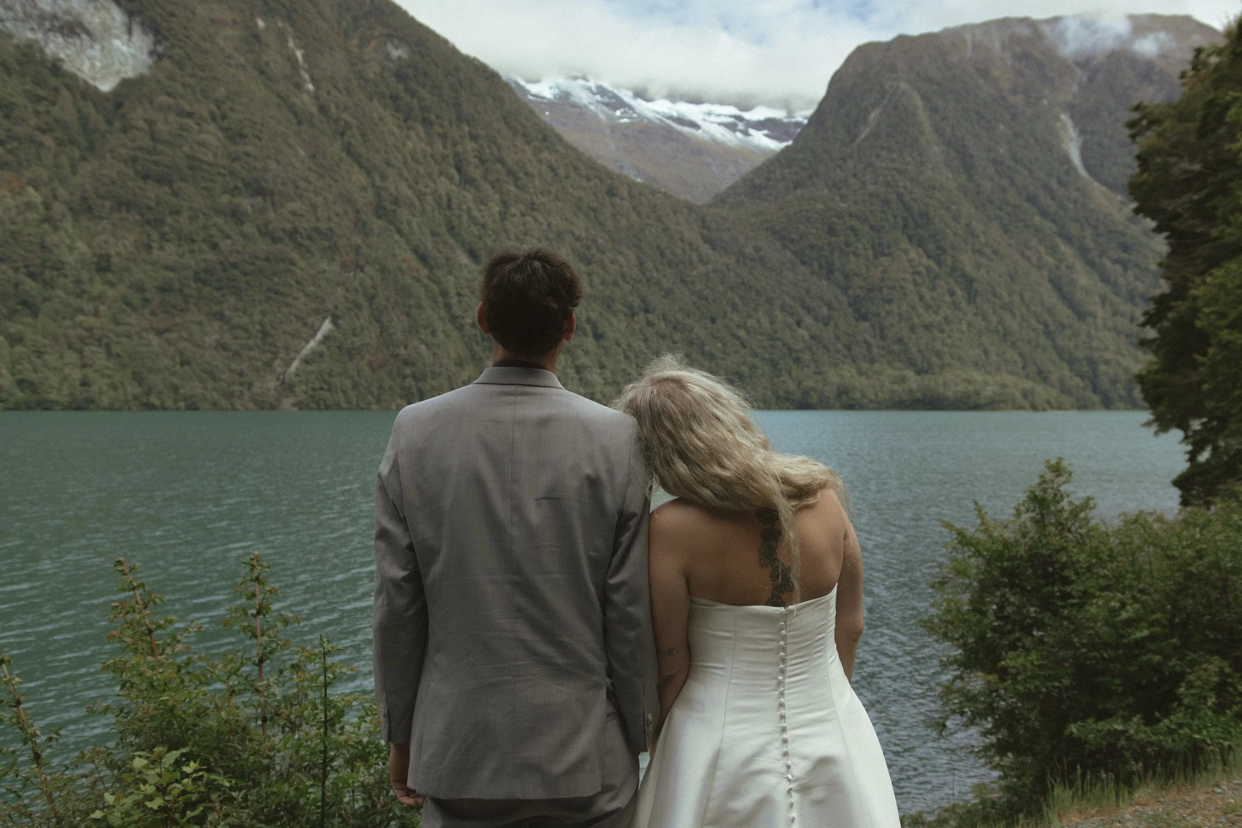 A couple in wedding attire standing by a lake with mountains in the background, seen from behind.