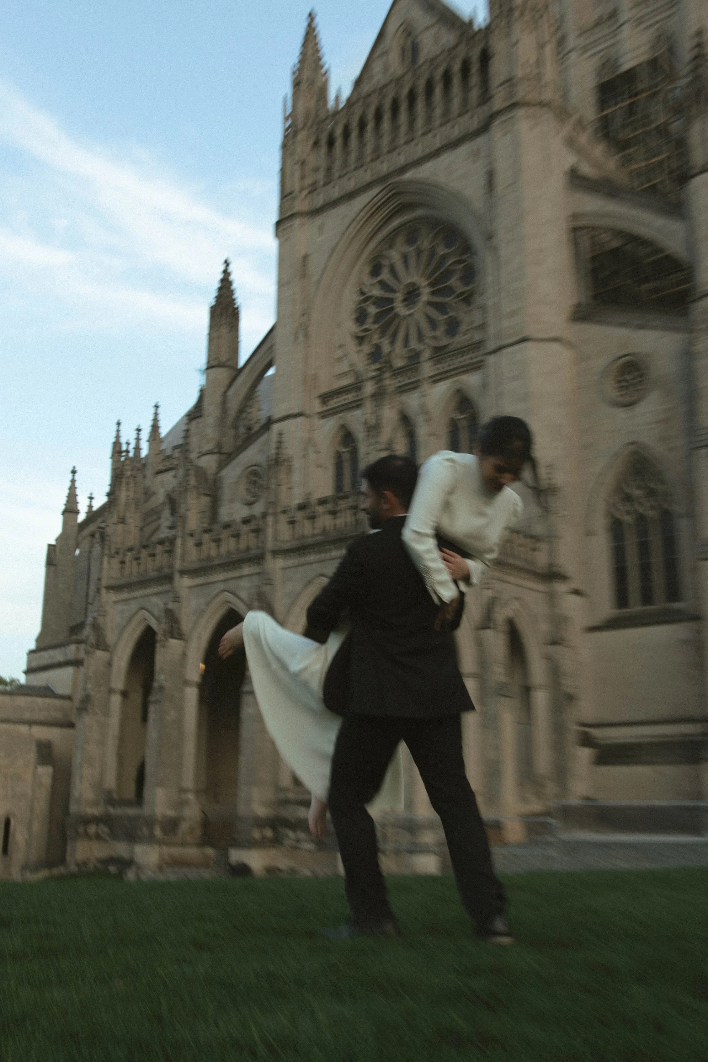 A man in a black suit carrying a woman in a white dress outside a large, historic cathedral during dusk.