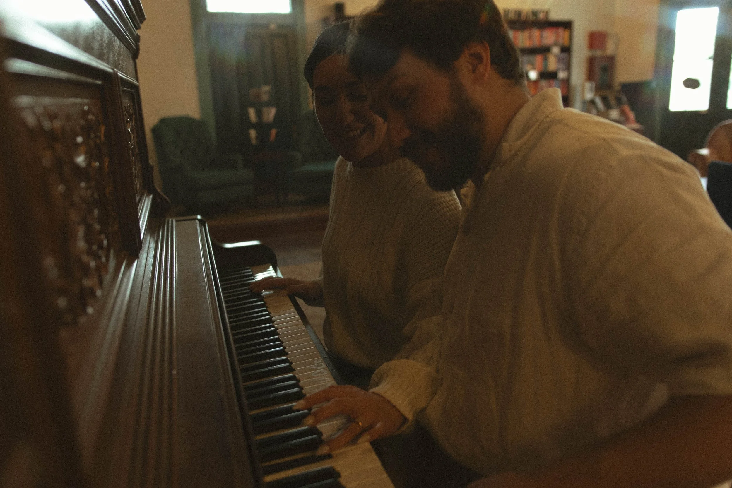 A man and woman sitting close together at a piano, smiling and playing music in a warmly lit room with bookshelves and green chairs in the background.