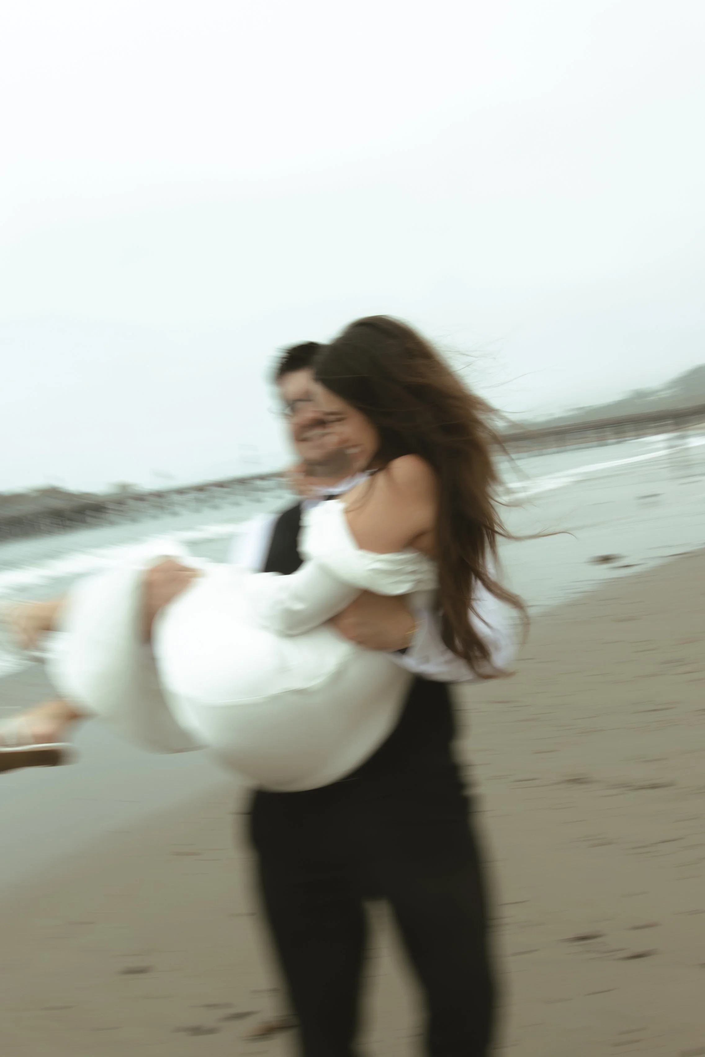 A woman in a white dress being carried by a man on a beach, both smiling with the ocean in the background.