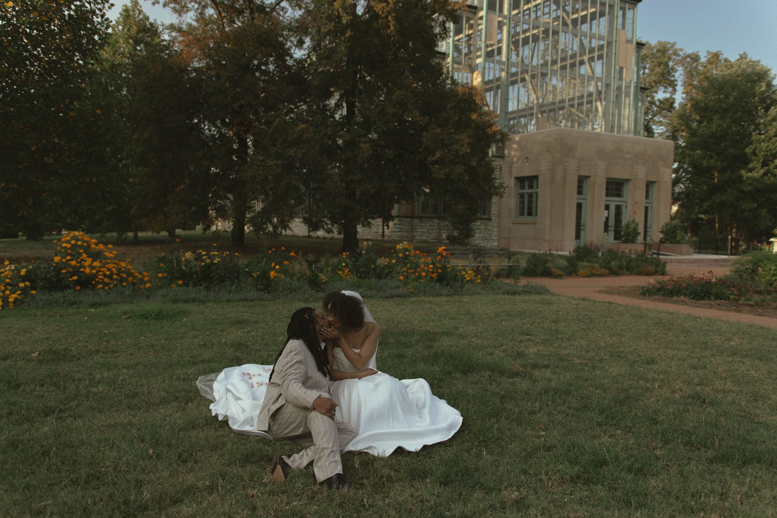 A couple dressed in wedding attire sitting on the grass kissing outdoors in front of a building with glass windows and surrounded by trees and flowers.