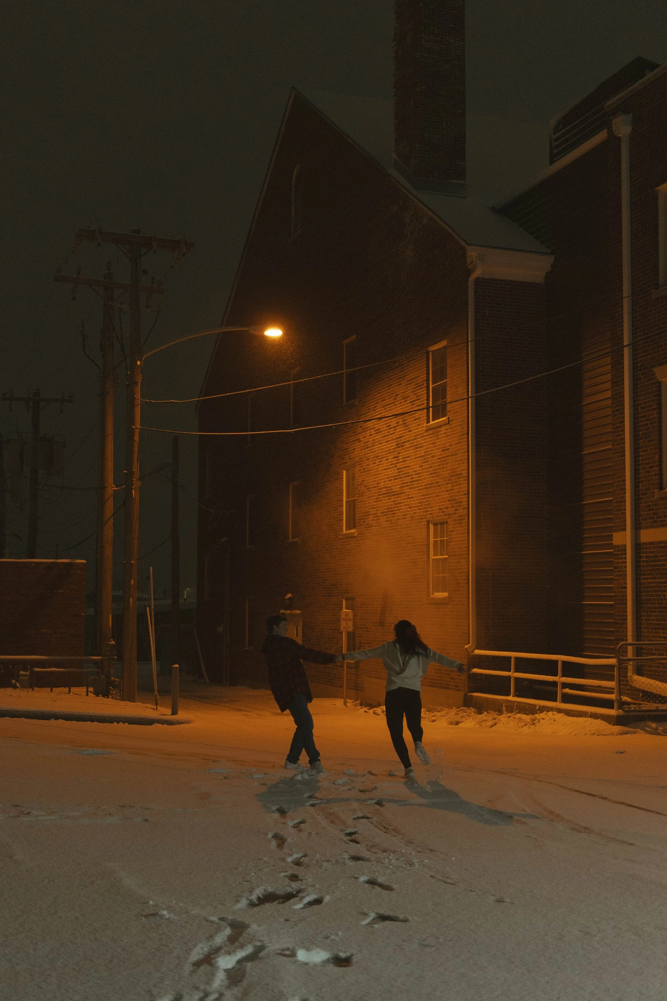 Two people hold hands and dance on a snow-covered street at night, illuminated by a nearby streetlight, with brick buildings and utility poles in the background.