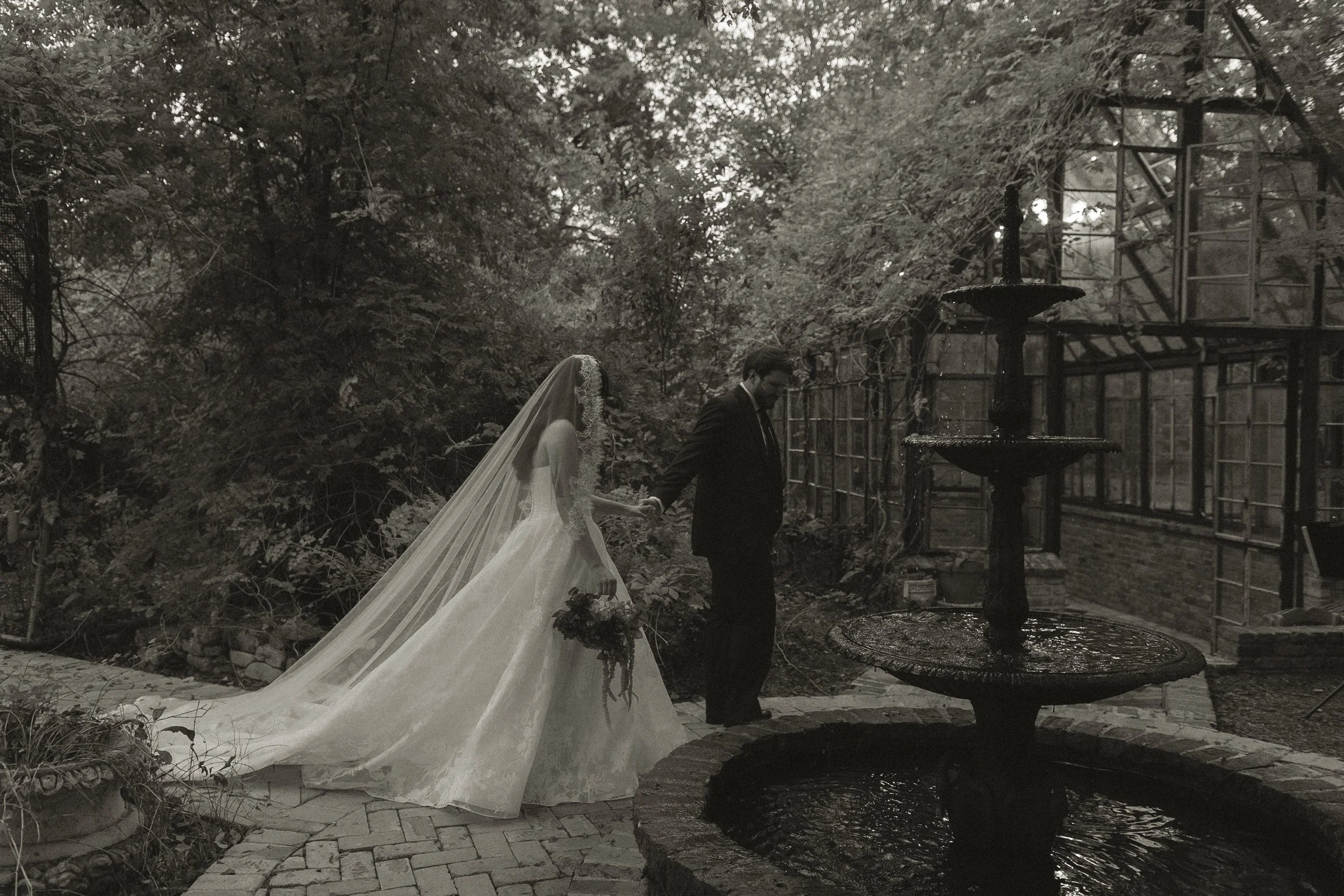 A bride in a wedding dress and veil holding a bouquet stands beside a man in a suit in a garden near a fountain, with lush trees and a greenhouse structure in the background.