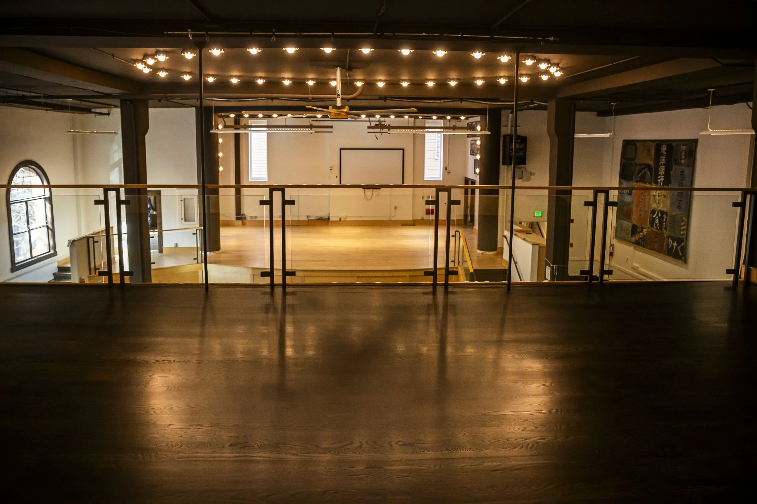 Empty urban dance studio with wooden floors, black ceiling, decorative lights, large windows, and a whiteboard at the back.