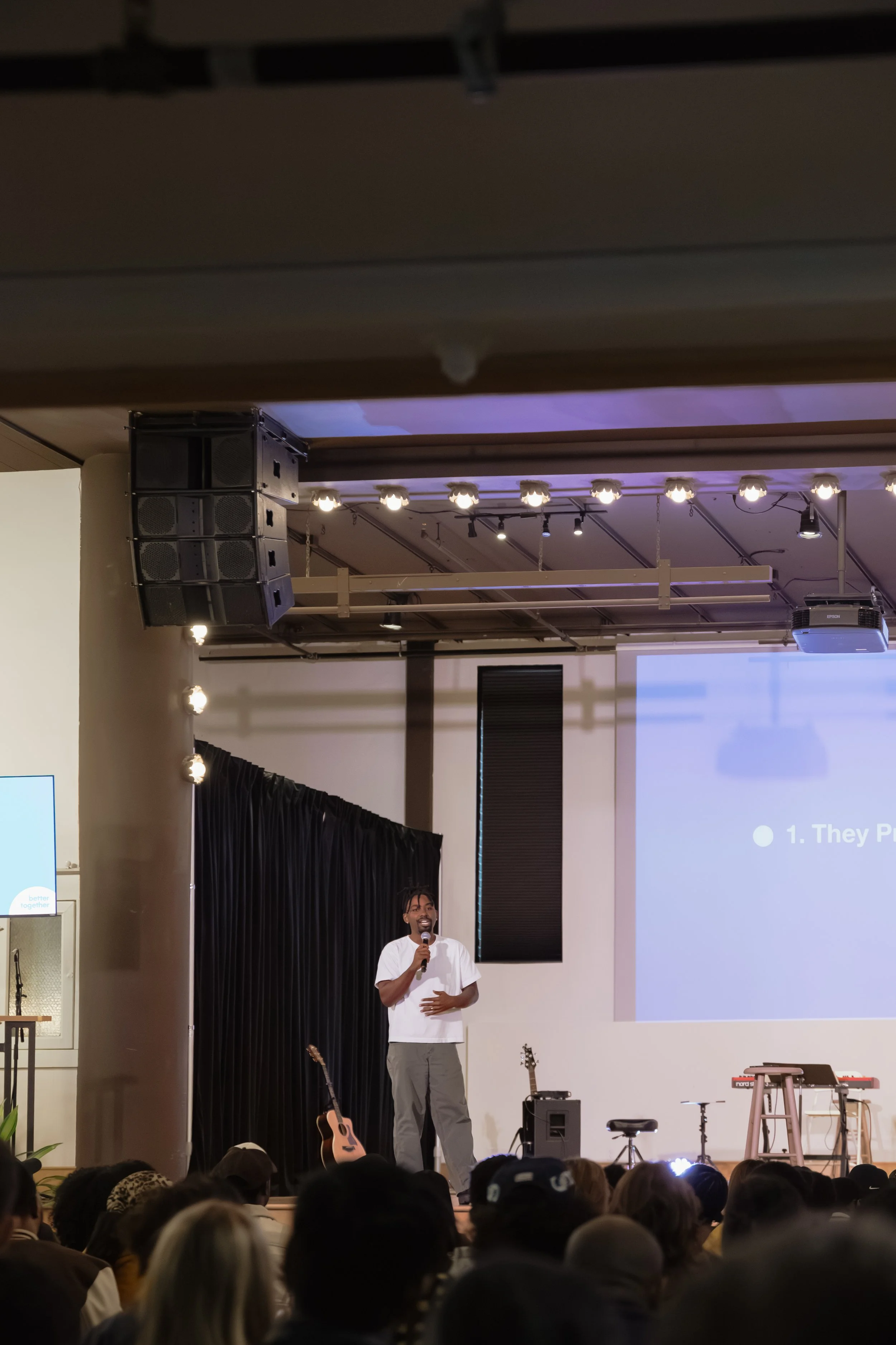 A man stands on stage holding a microphone, speaking to an audience. There are musical instruments such as a guitar and keyboard on stage, with a large screen behind him displaying a presentation.