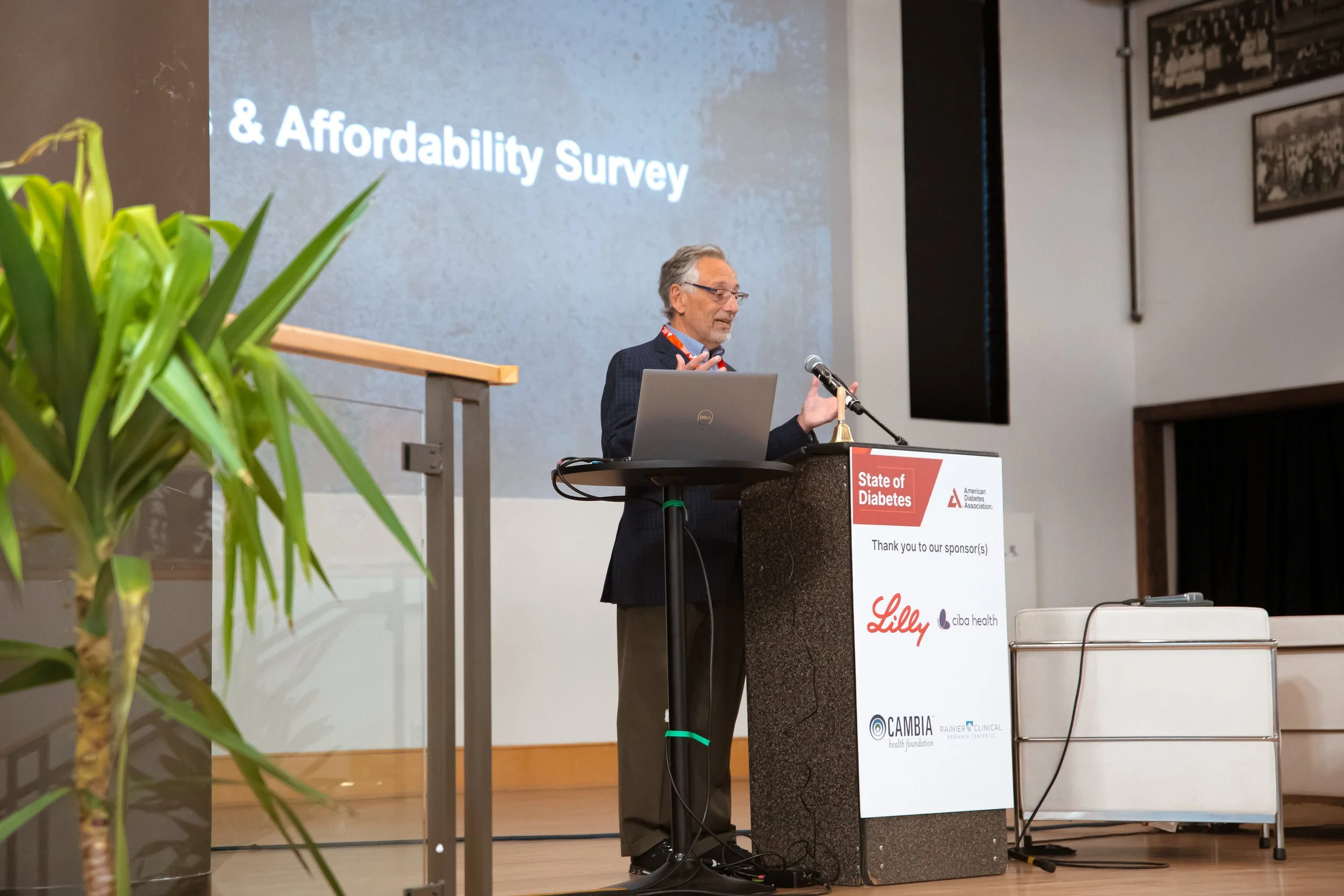 A man with glasses and gray hair giving a presentation at a conference, standing behind a podium with a sign that reads 'State of Diabetes.' There is a large screen behind him displaying text and a laptop in front of him on the podium.