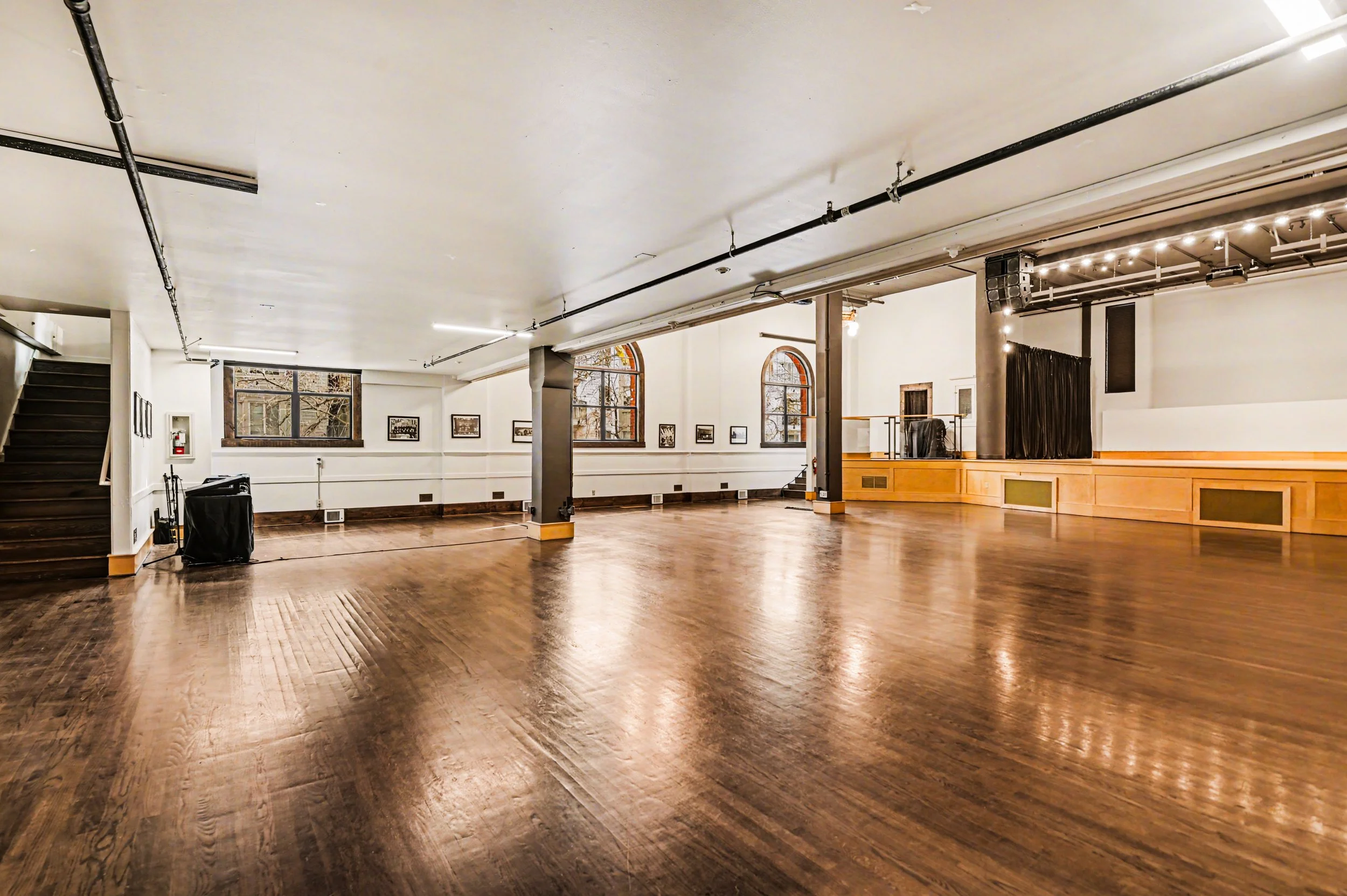 Nippon kan theatre, seattle, washington, event space, wedding,An empty dance or event hall with polished wooden flooring, white walls, large arched windows, and a small stage area with black curtains, decorated with string lights.