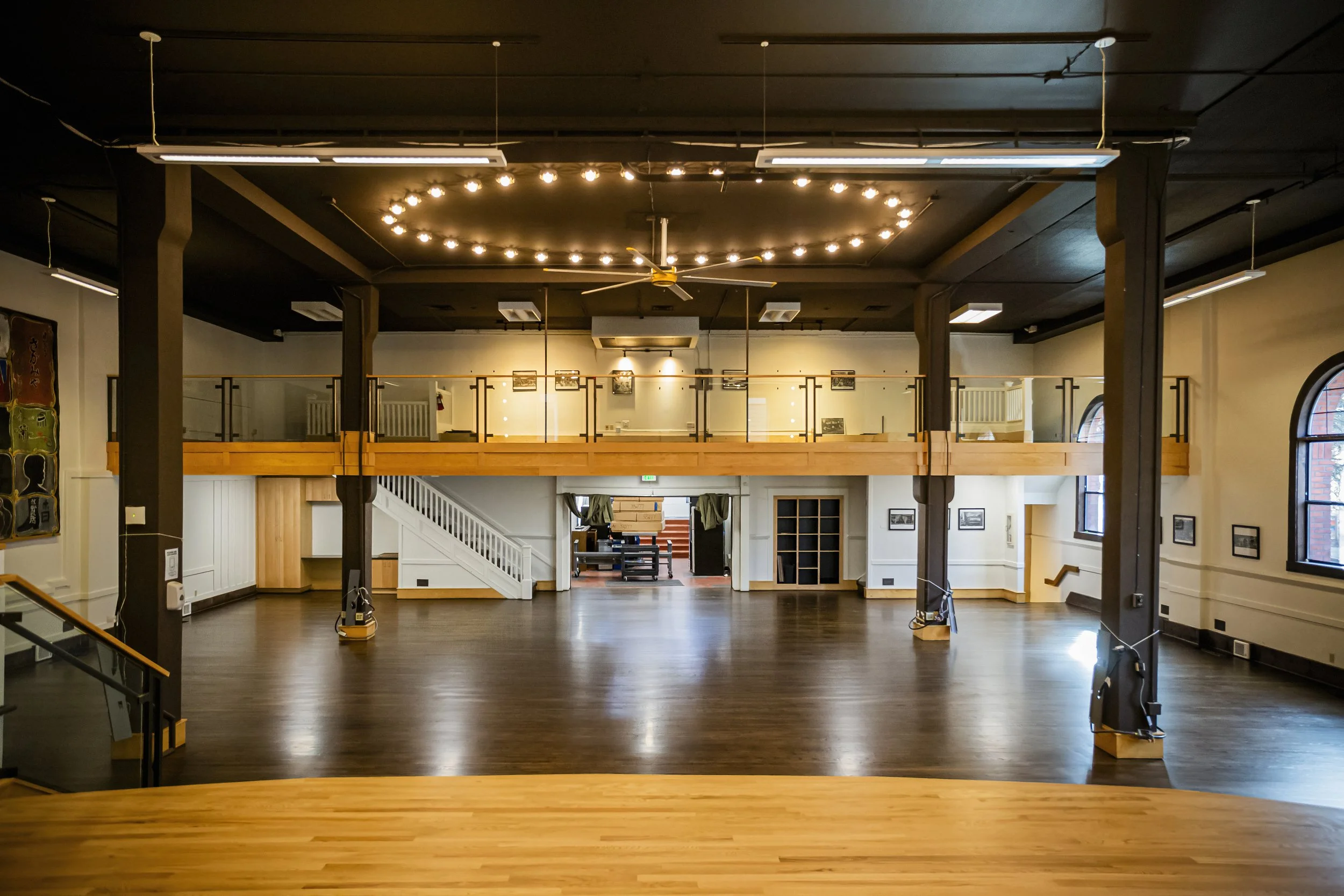 Empty interior of a large, two-story open space with wooden flooring, black ceiling, and large windows, featuring a balcony railing on the upper level and framed photos on the walls.