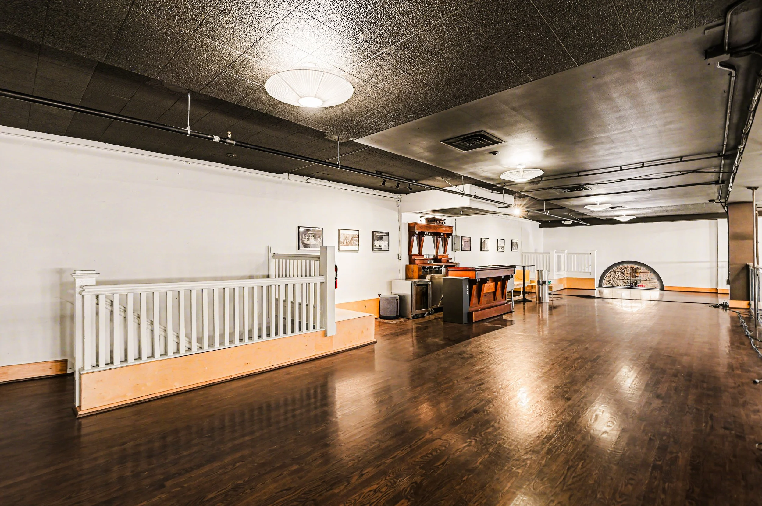 Nippon kan theatre, seaAn empty dance hall or event space with dark wooden floors, white walls decorated with framed pictures, and a low stage area with railings. There are ceiling lights and a small stage or platform with liquor bottles and a piano.