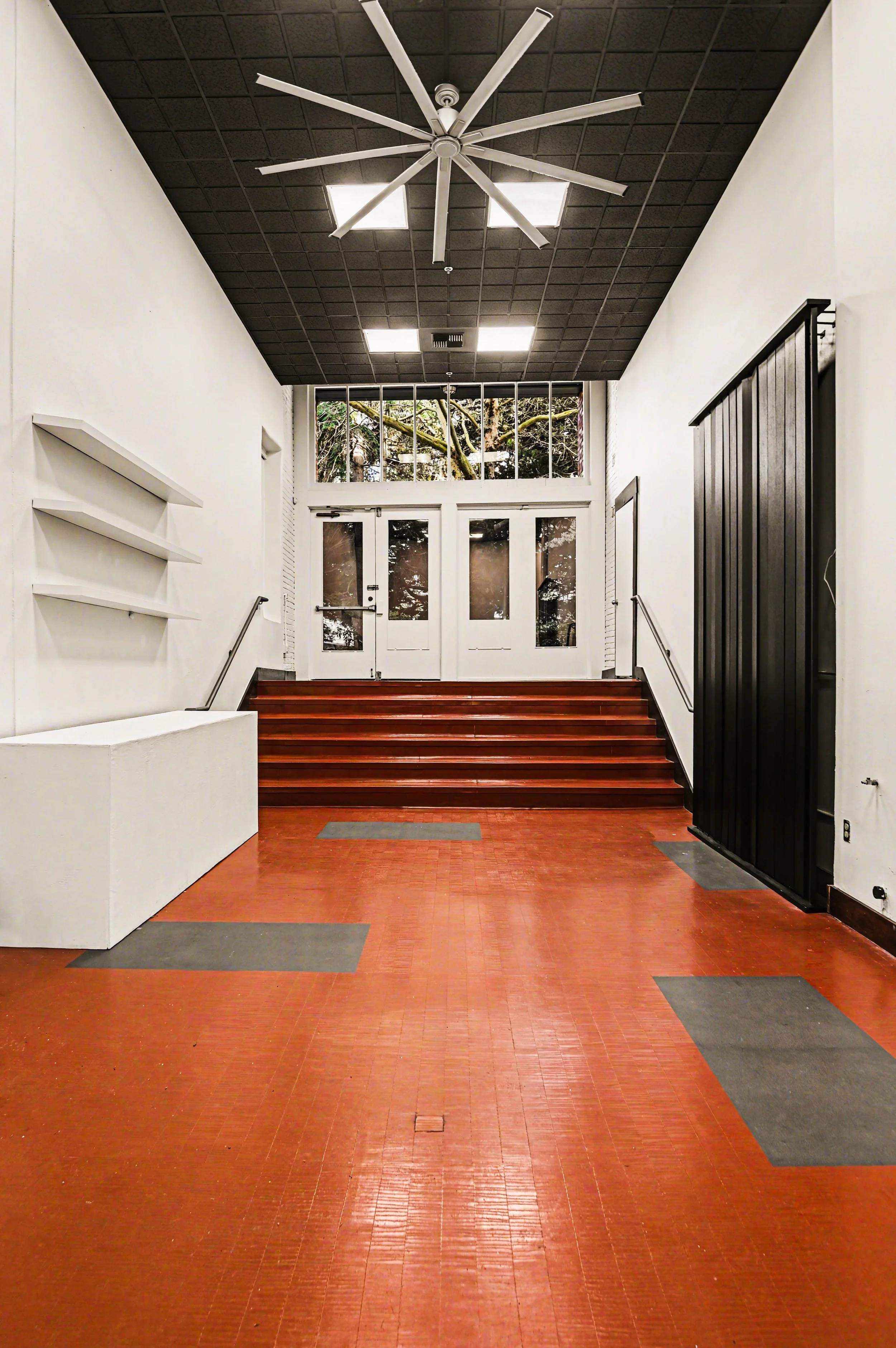 Nippon kan theatre, seattle, washington, event space, Interior of a building entryway with red tile floor, white walls, black ceiling, a large white ceiling fan, and glass double doors leading outside, with stairs and trees visible through the glass.