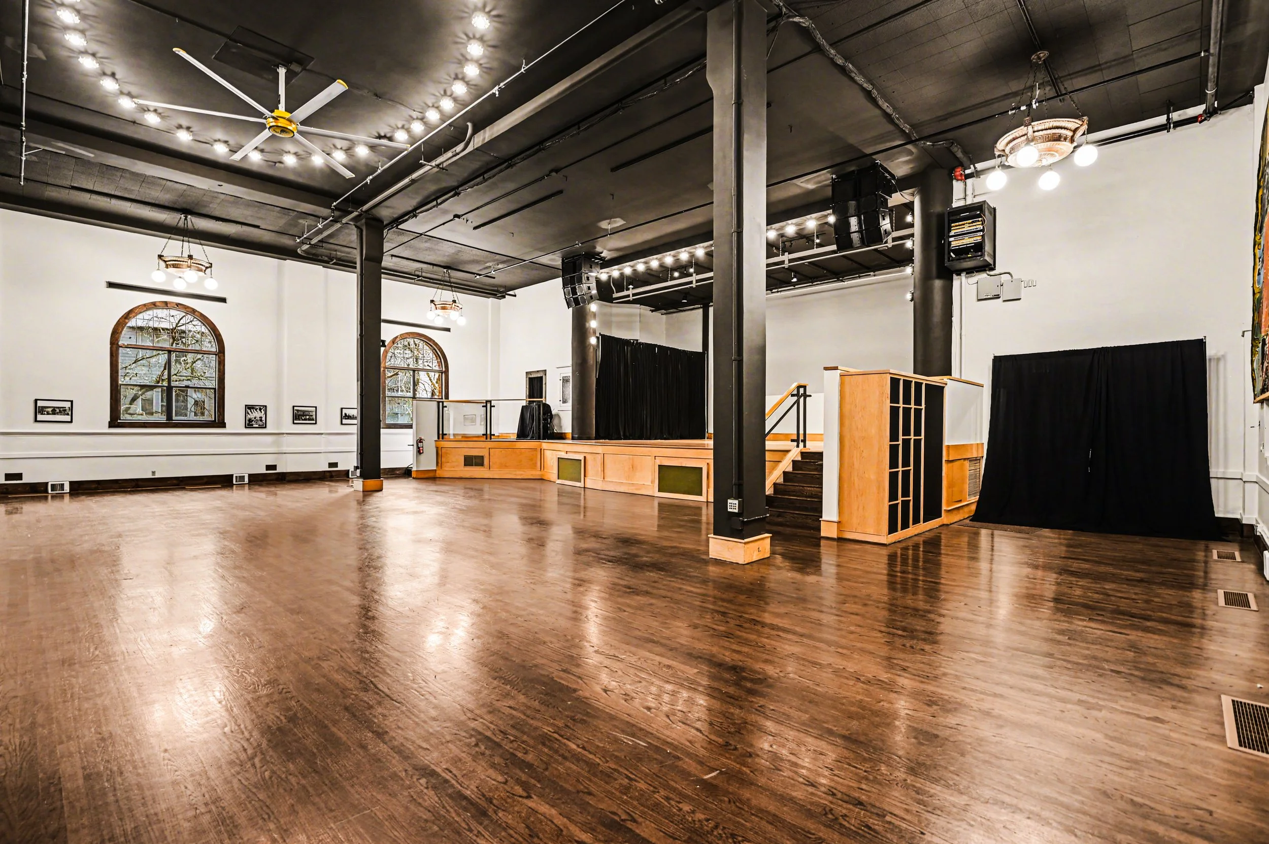 Nippon kan theatre, seattle, washington, event space, wedding,Empty event hall with hardwood floors, white walls, arched windows, stage with black curtains, and black ceiling with spotlights and ceiling fans.